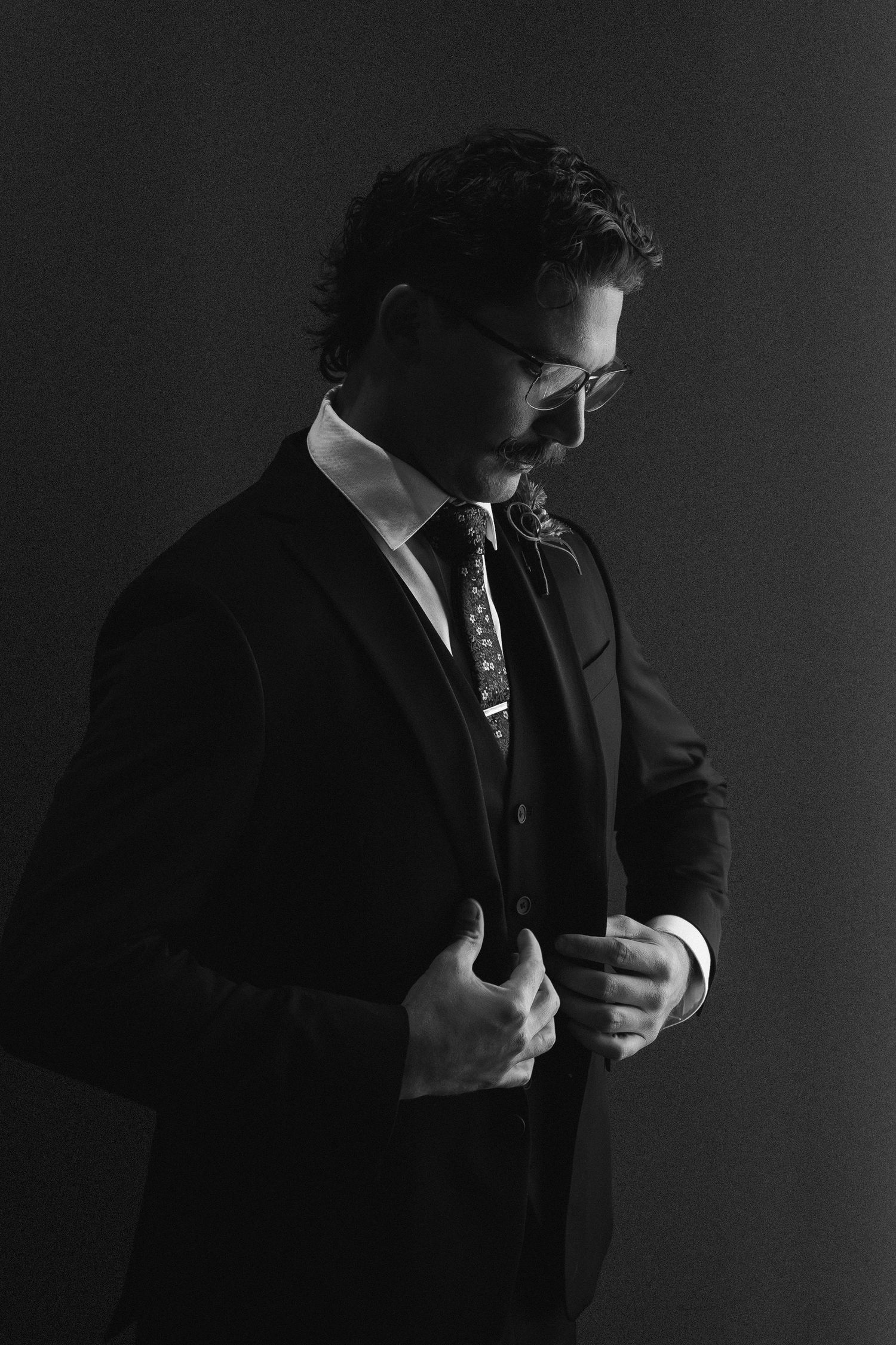 Dapper groom in black suit and tie adjusting jacket lapels in dramatic black and white portrait.