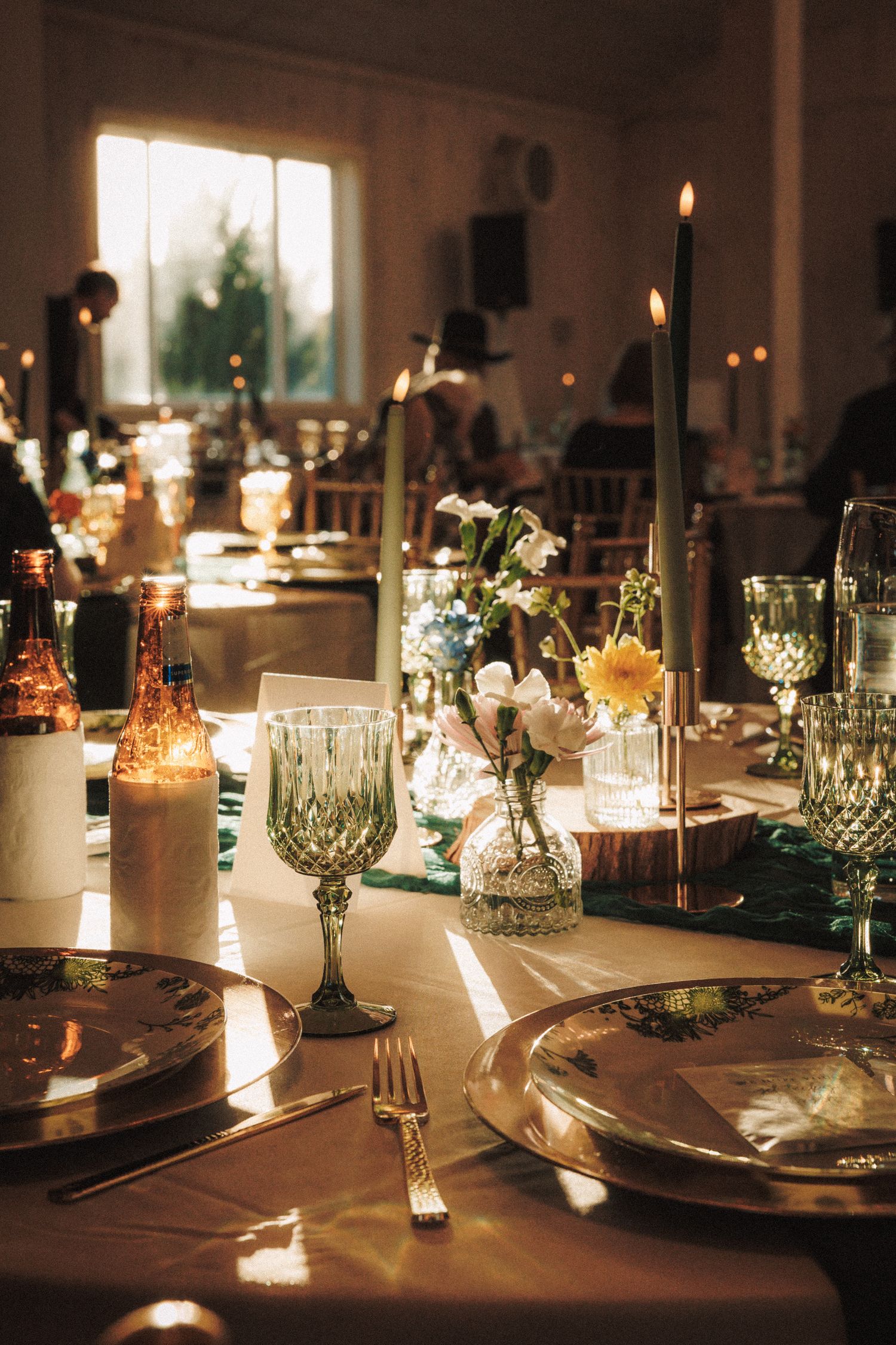 Elegant wedding reception table with green glassware, candles, and floral centerpieces in warm light.