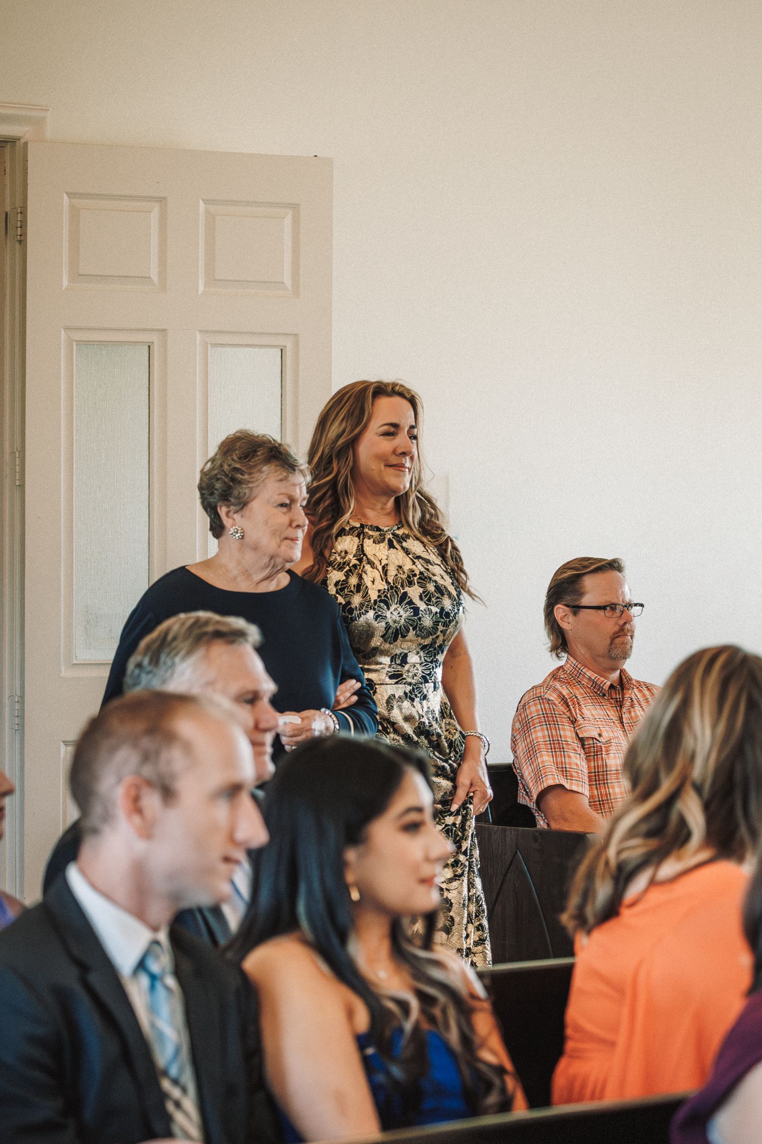 Wedding guests smile and mingle inside bright white ceremony venue.