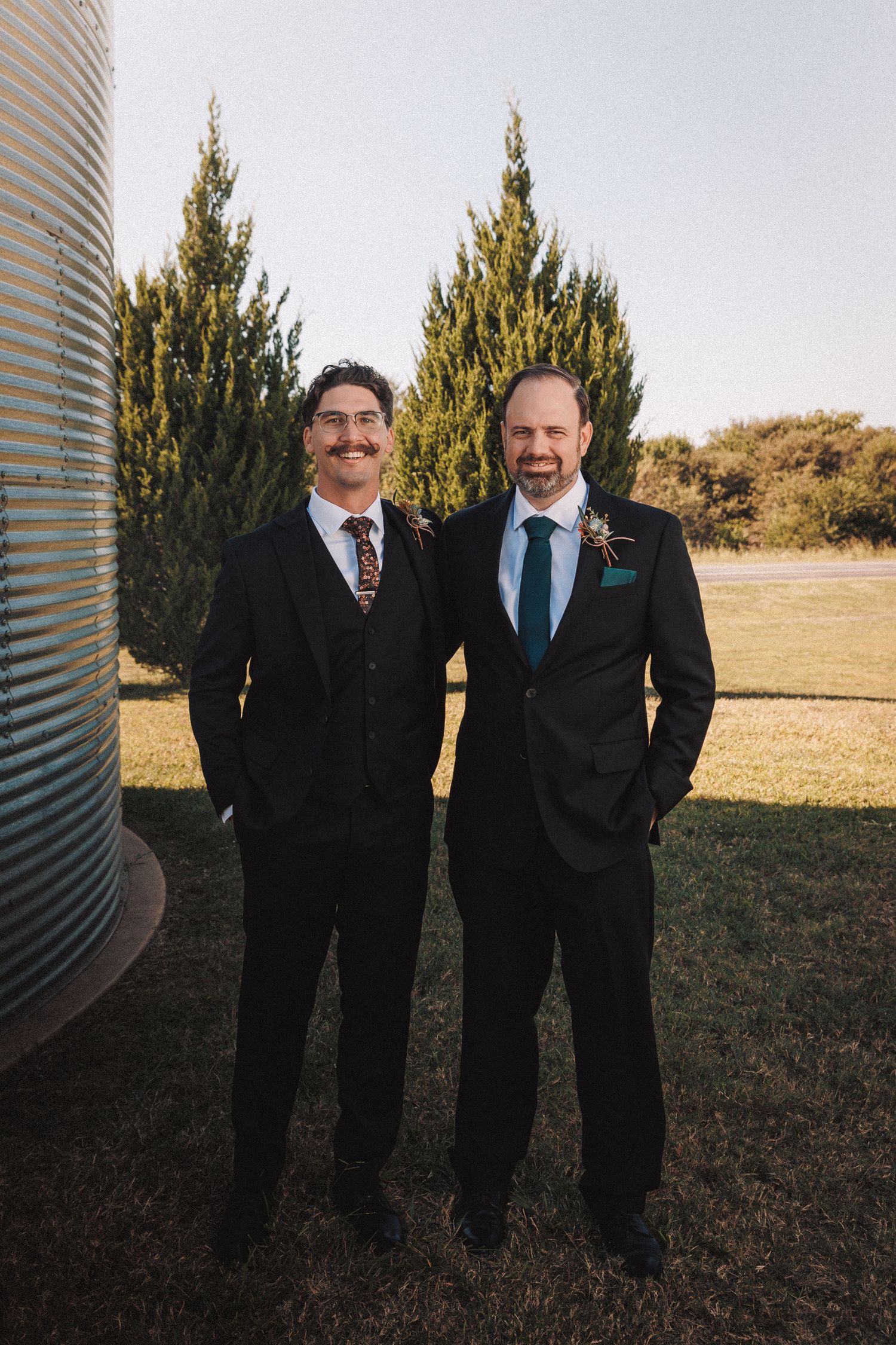 Two grooms in black suits smile together outdoors near evergreen trees at their wedding.