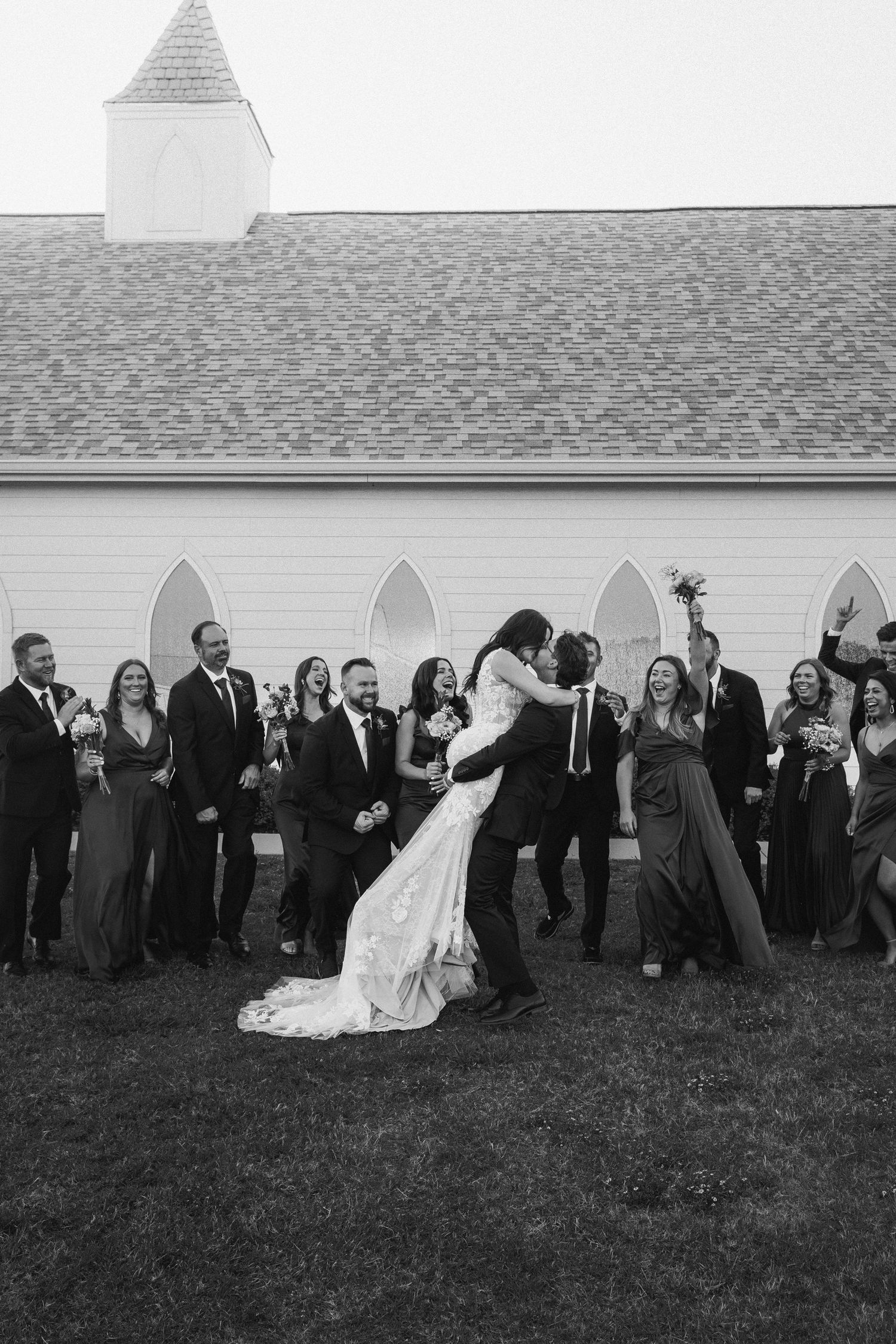 Wedding party celebrates outside a white chapel as groom lifts bride in a kiss.