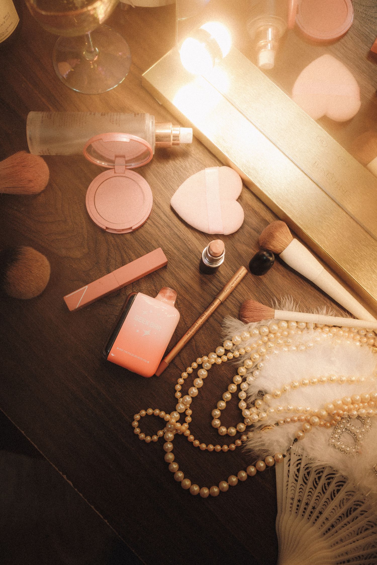 Pink makeup products, pearl necklace, and feather fan arranged on a wooden vanity table with warm glowing mirror lights.