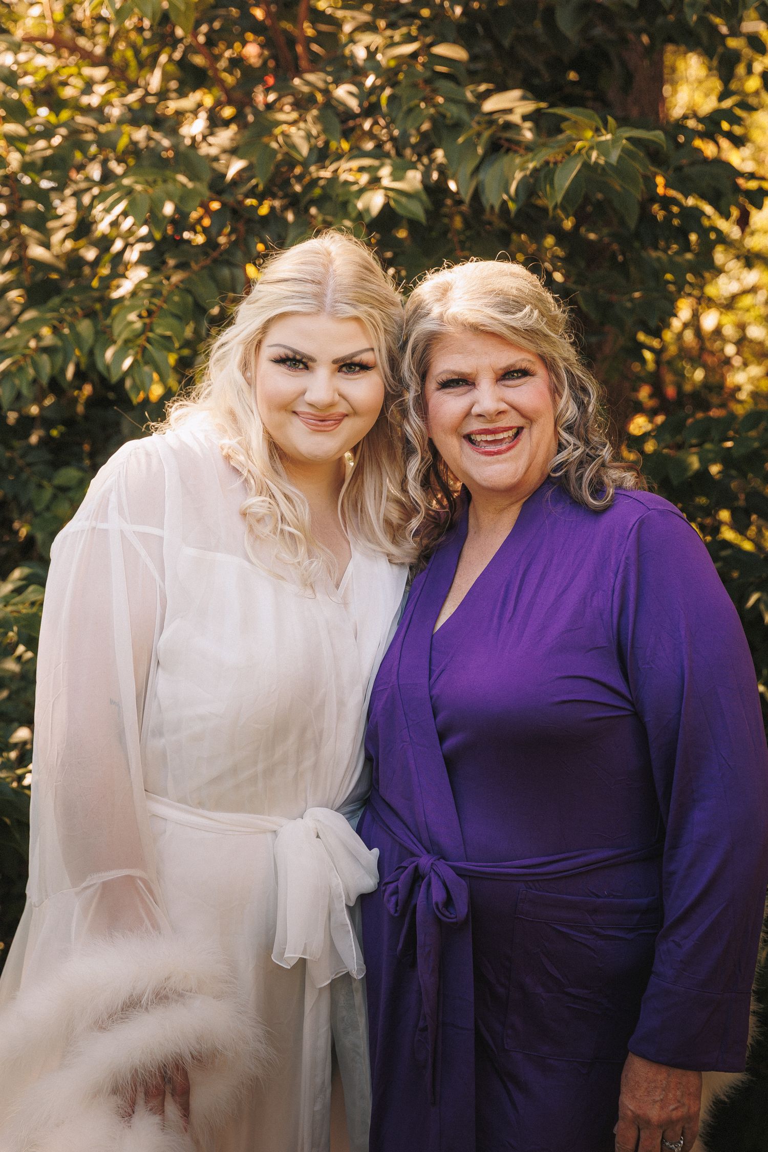 Bride in sheer white robe posing with woman in purple dress outdoors in golden autumn light.