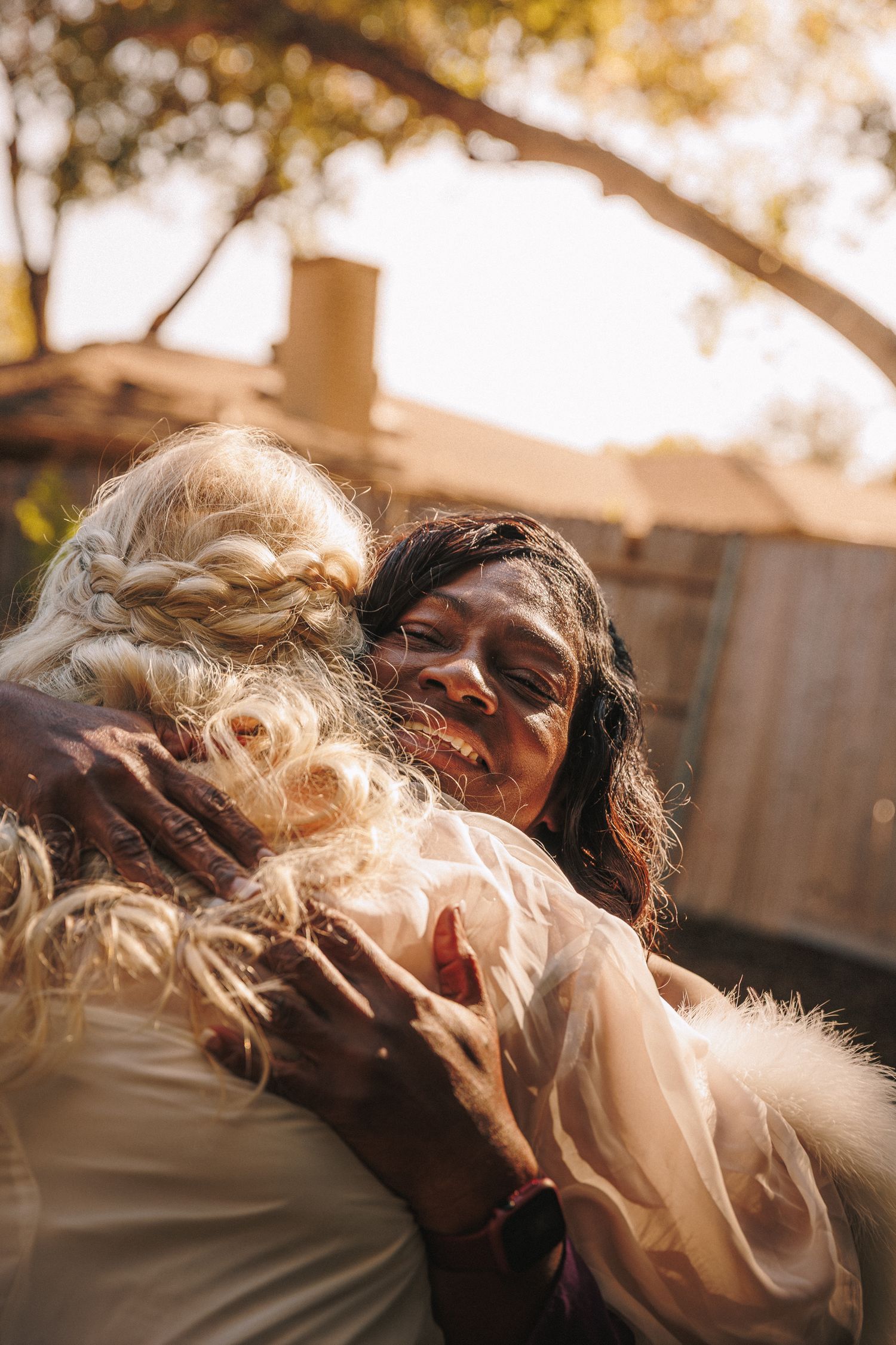 Emotional wedding day hug between bride in white gown and guest outdoors in golden sunlight.