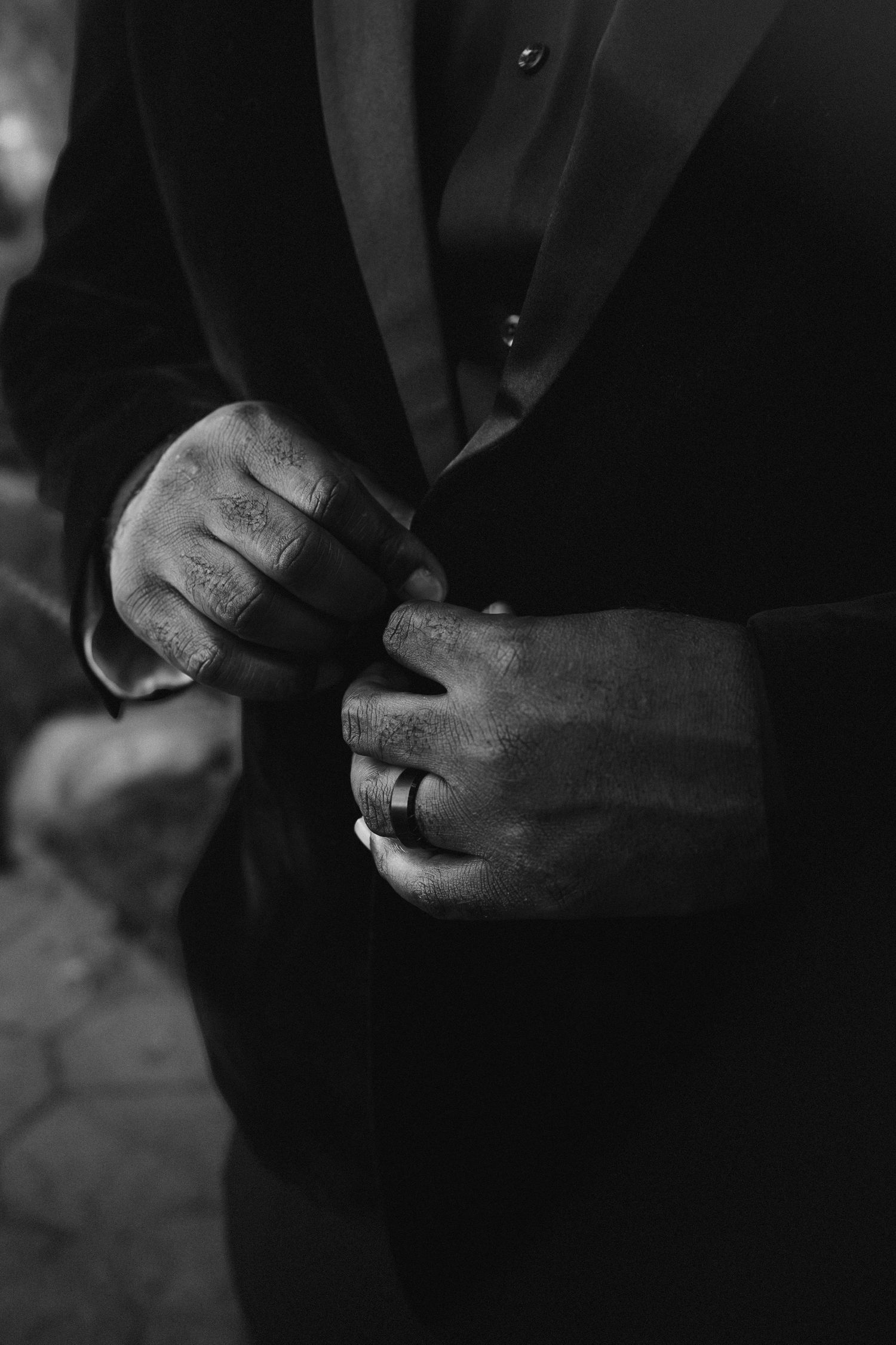 Black and white close-up of groom adjusting tuxedo jacket with tattooed hands and wedding ring.