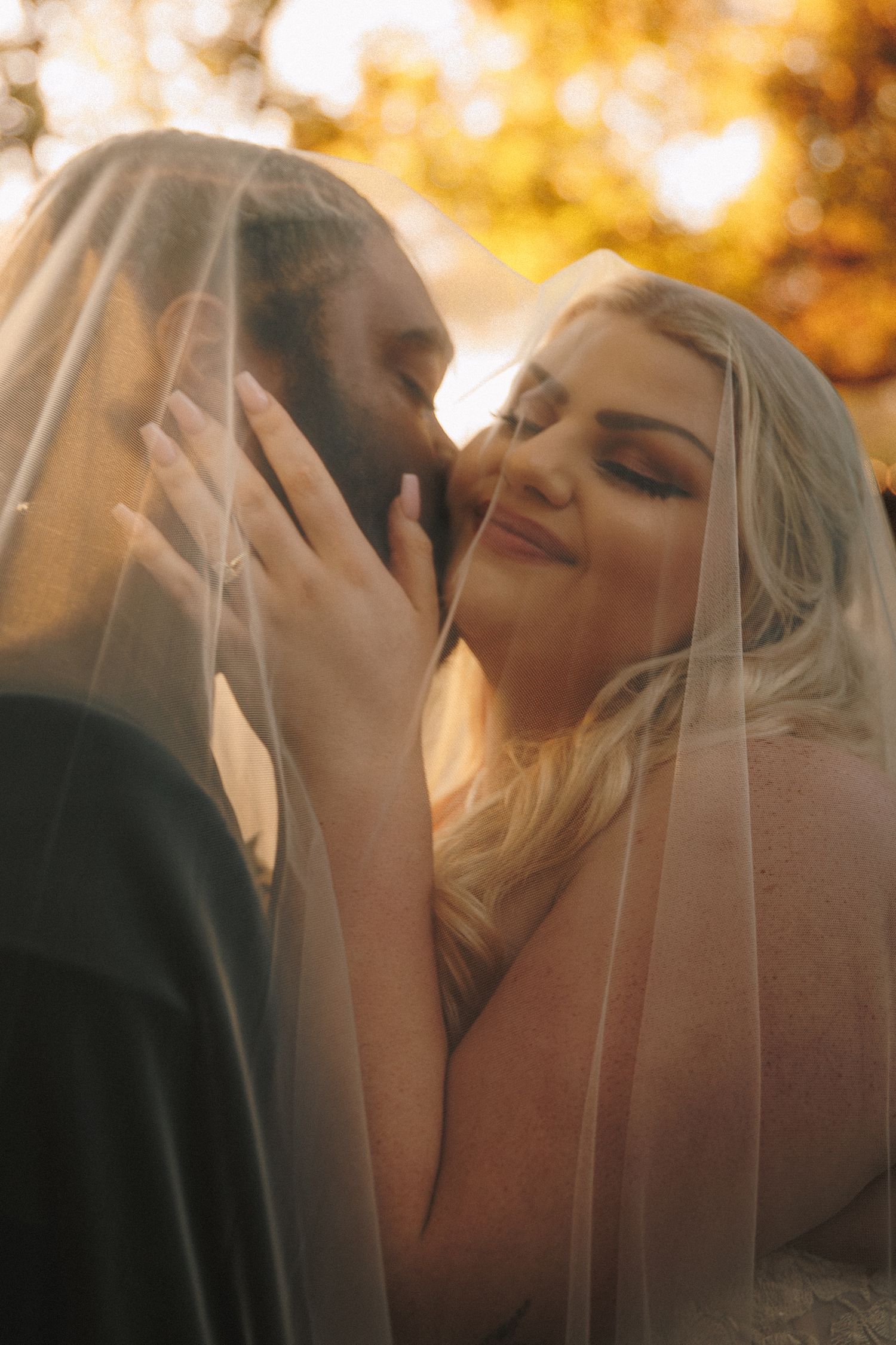 Bride and groom share a tender moment together under a flowing wedding veil in golden light.