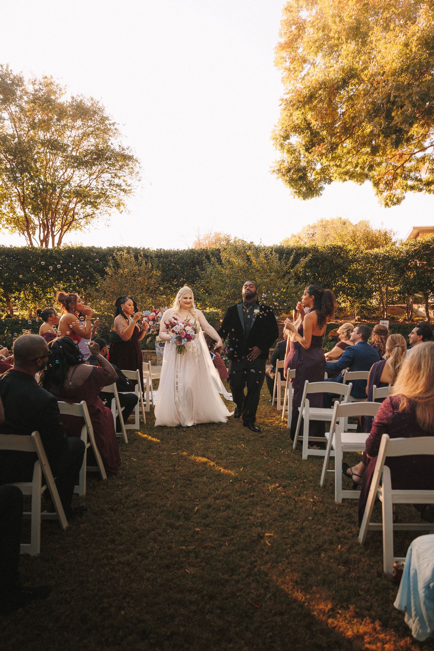 Newlywed couple walks down the aisle at sunset outdoor wedding ceremony as guests celebrate with confetti.