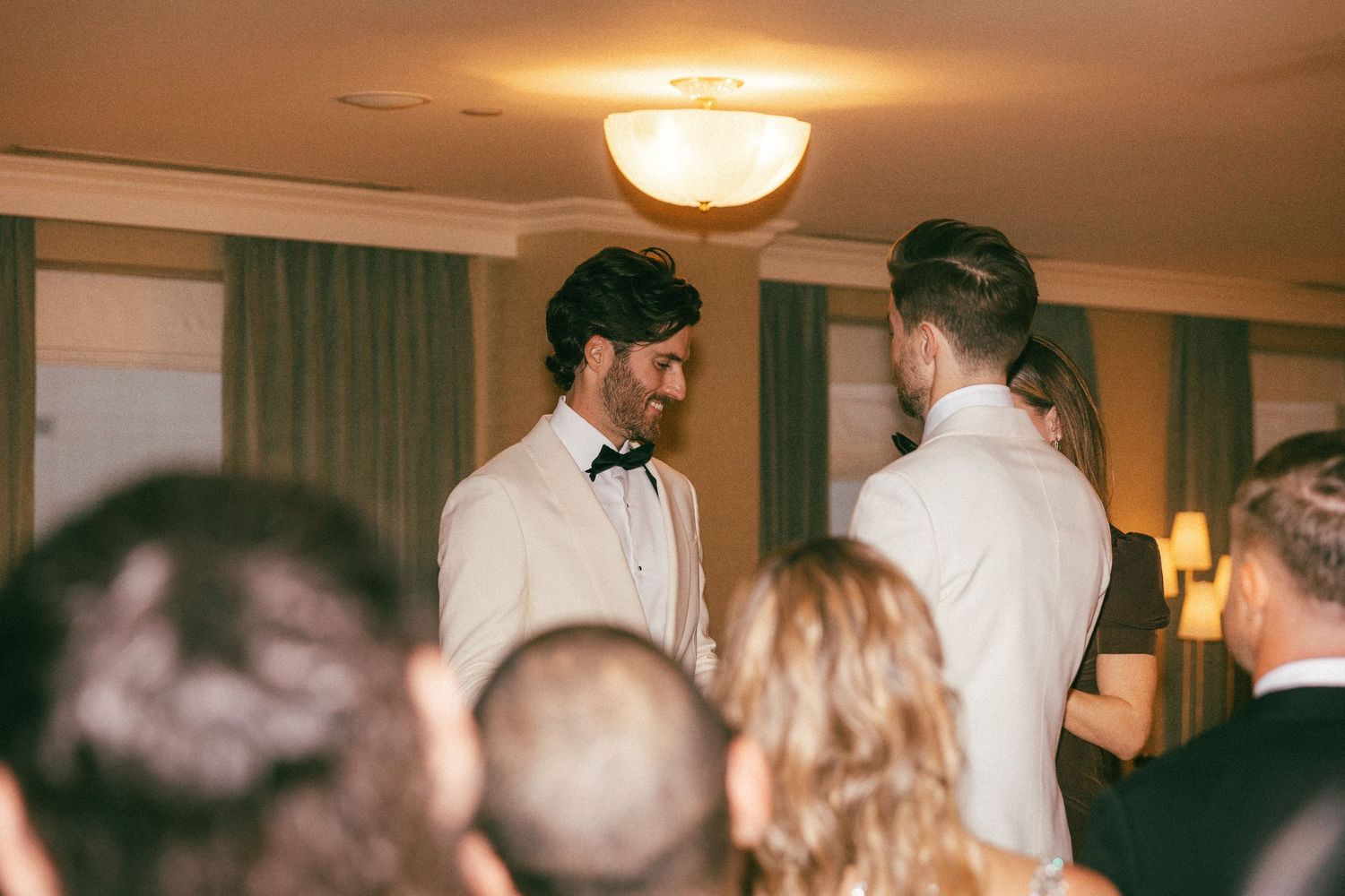 Groom in white tuxedo smiling warmly at partner during intimate indoor wedding ceremony.