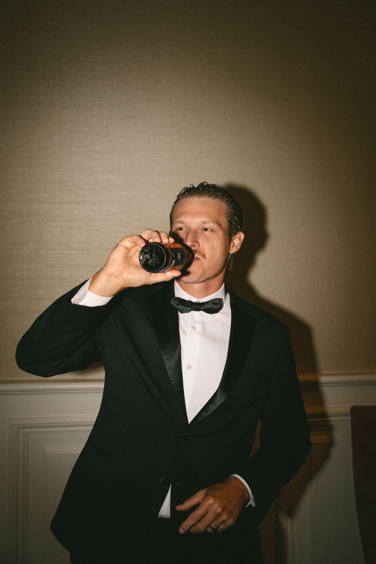 Groomsman in black tuxedo drinking from a small bottle at an elegant wedding reception venue.