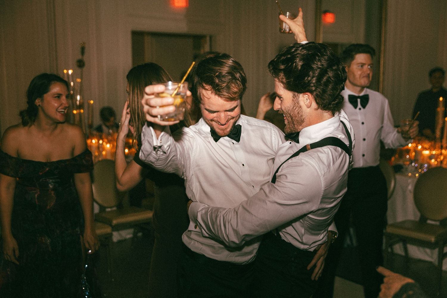 Two men dance energetically holding cocktails at a lively wedding reception.