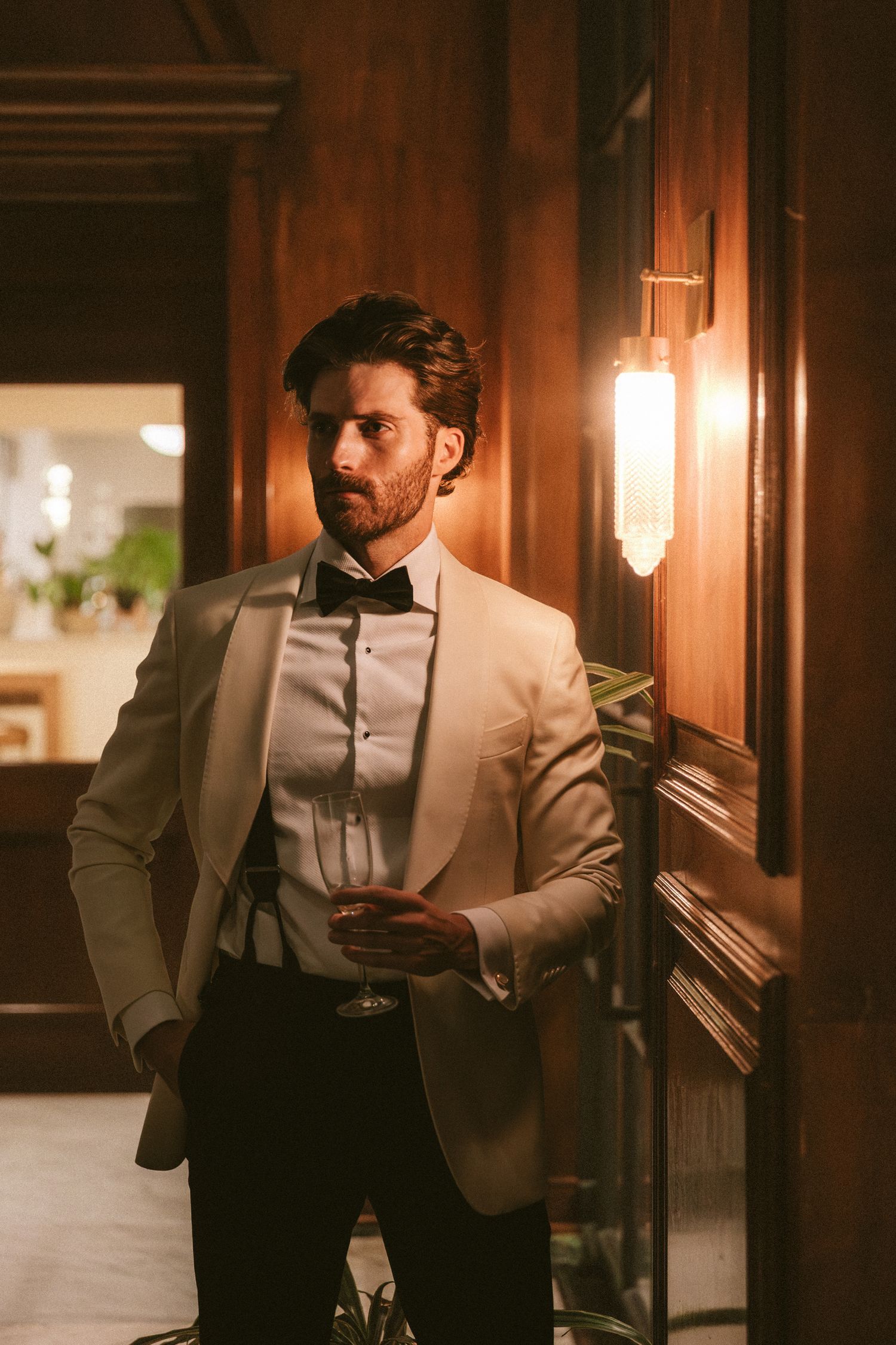 Stylish groom in ivory tuxedo jacket and black bow tie leans against wooden wall holding a drink.