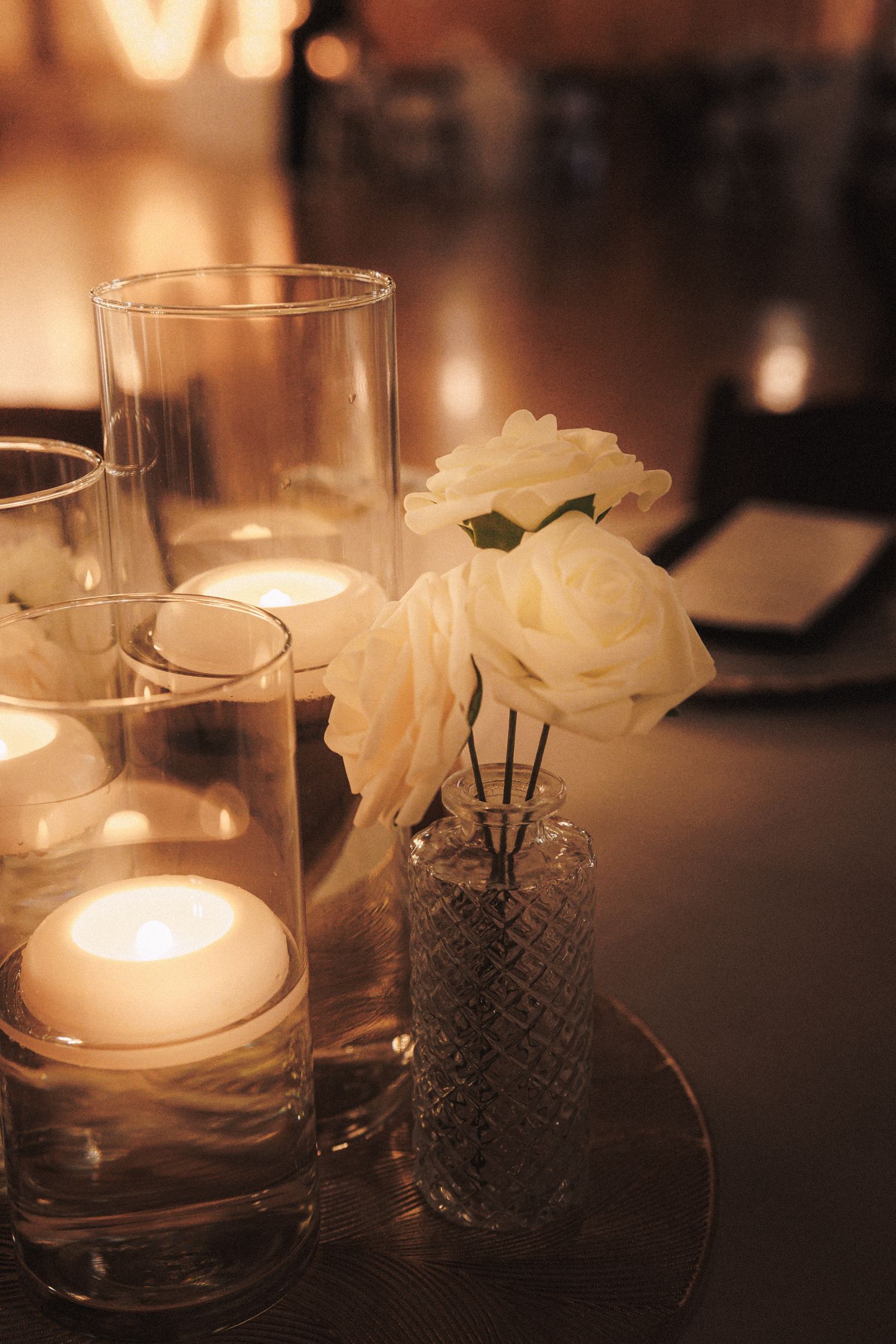 Elegant wedding centerpiece with glowing candles in glass cylinders and white roses in a crystal vase on a dark table.