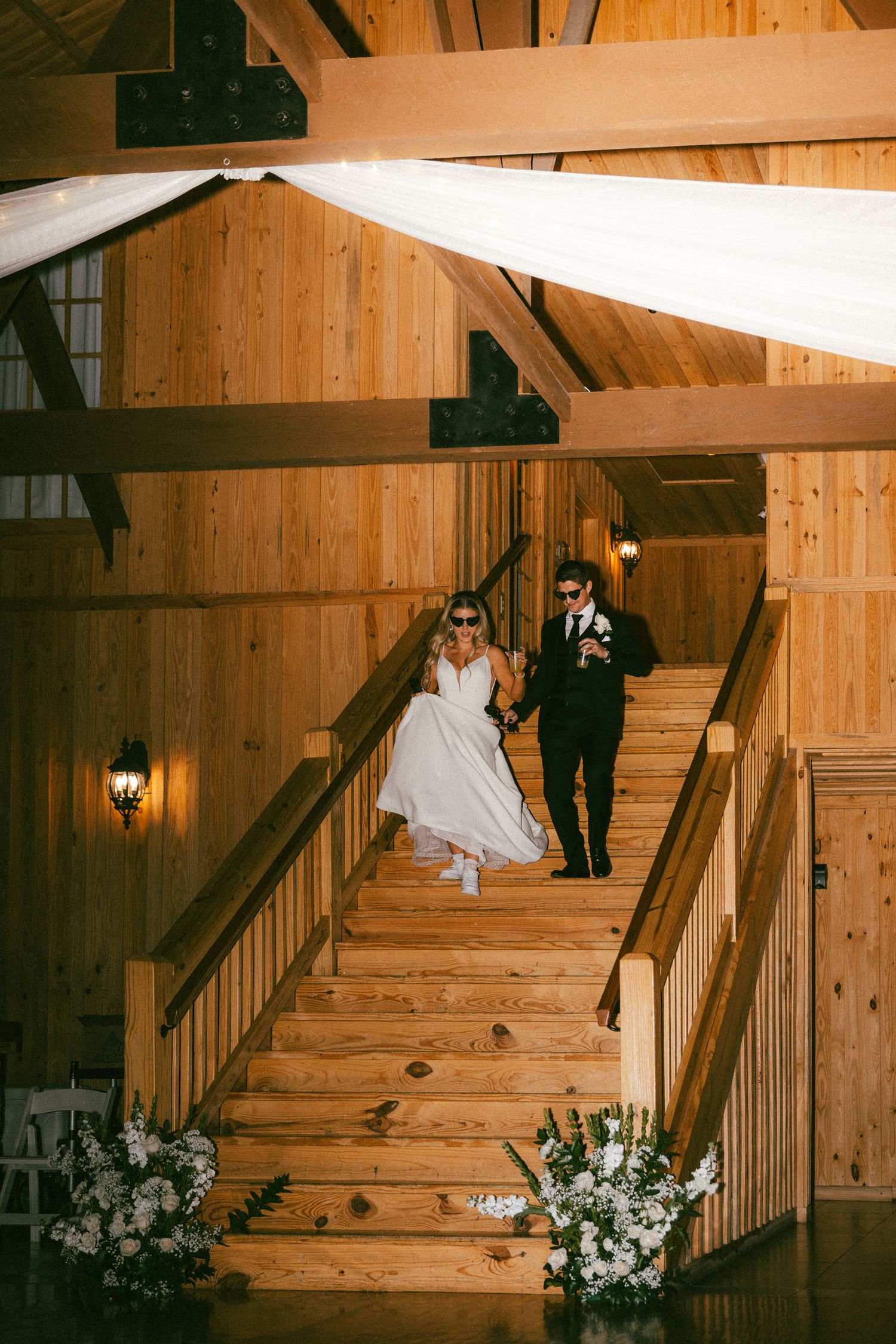 Bride and groom descend wooden staircase in rustic barn venue wearing fun sunglasses at reception.