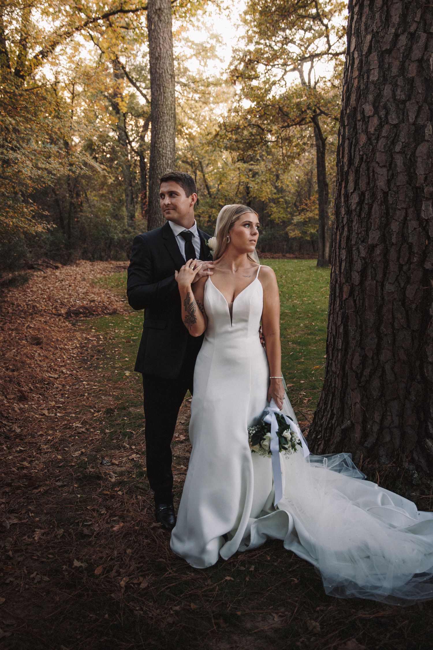 A bride and groom pose elegantly beside a large tree in an autumn forest setting.