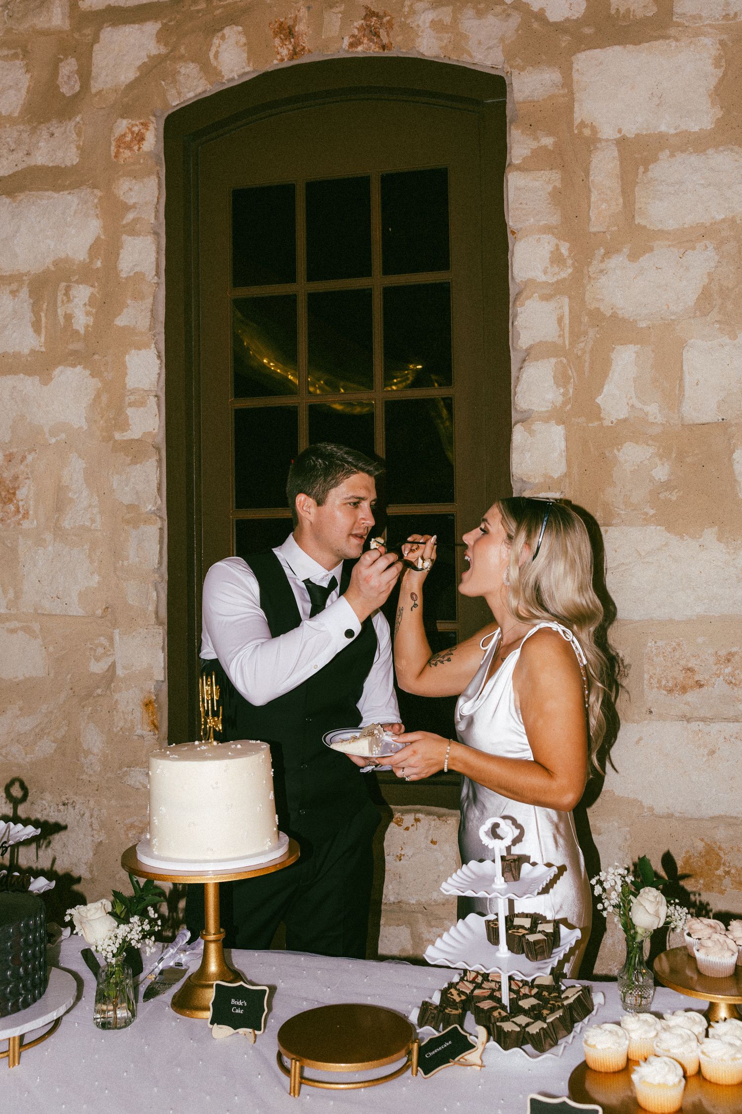 A bride and groom feed each other wedding cake at a dessert table against a rustic stone wall at their reception.