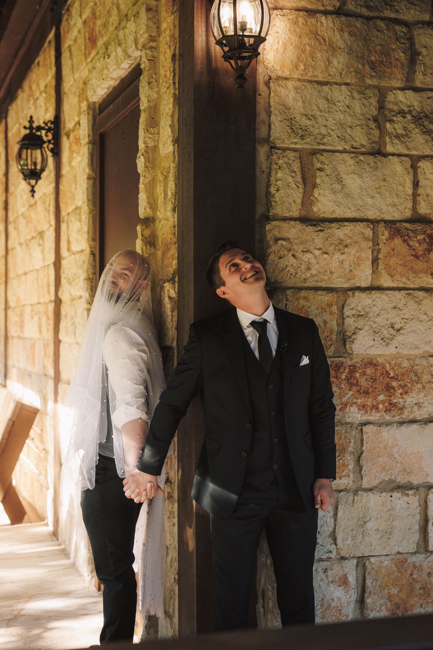 A bride and groom hold hands laughing together against a stone wall venue.