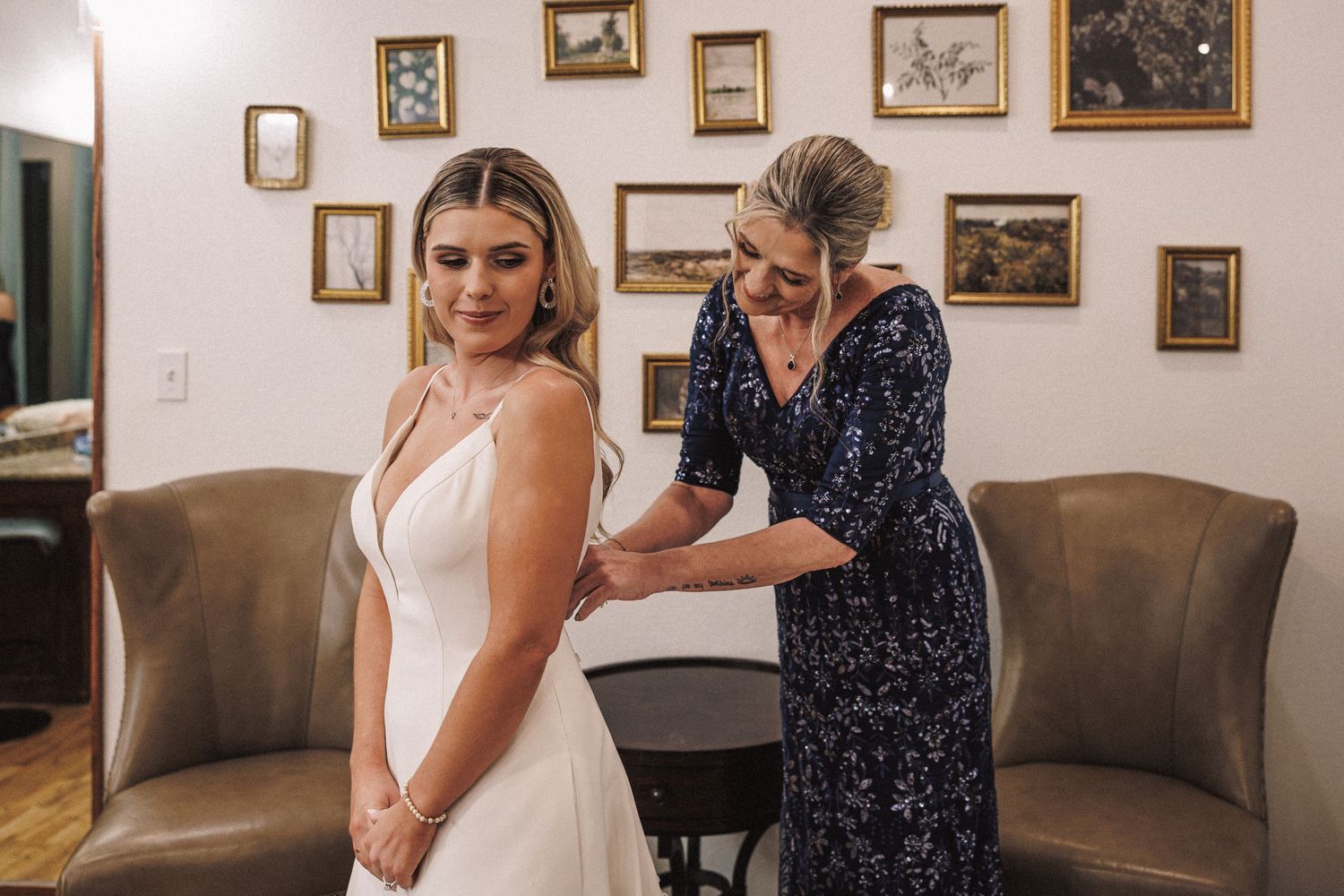 Smiling bride in white gown being helped into her dress by older woman in navy floral dress in elegant room.