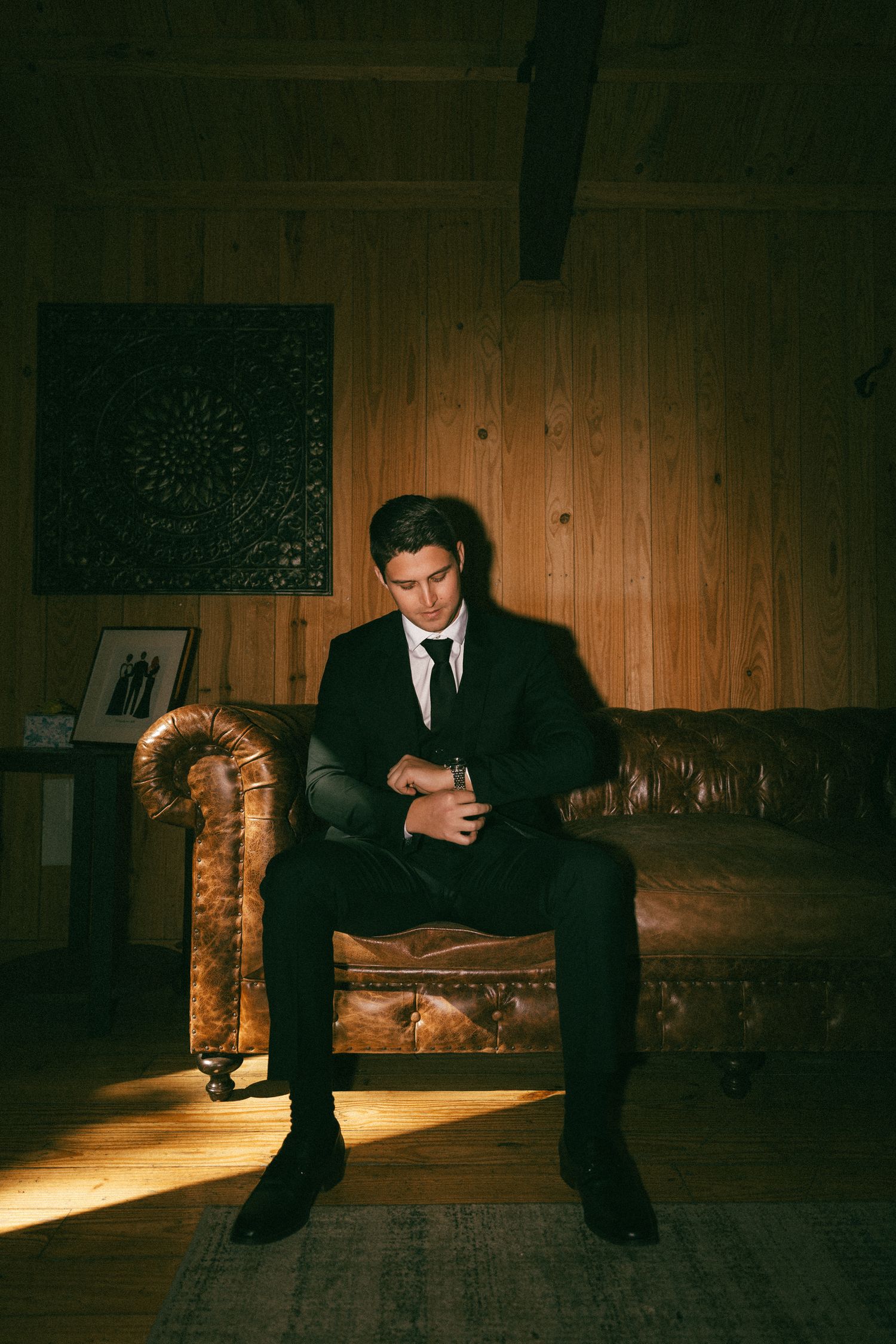 Groom in black suit sitting on leather sofa in warmly lit wooden room on wedding day.