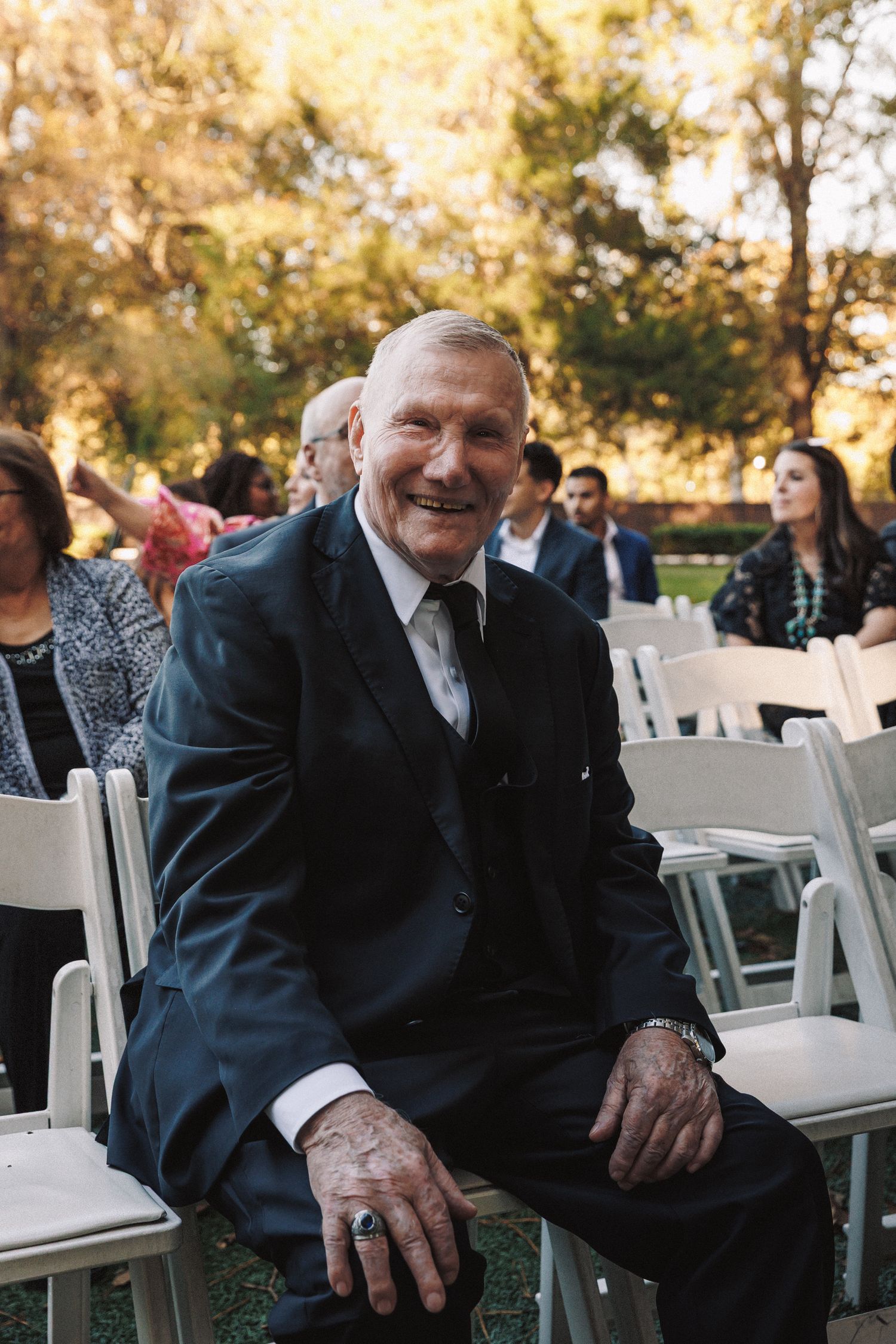 Smiling elderly wedding guest in dark suit seated in white chair at an outdoor ceremony.