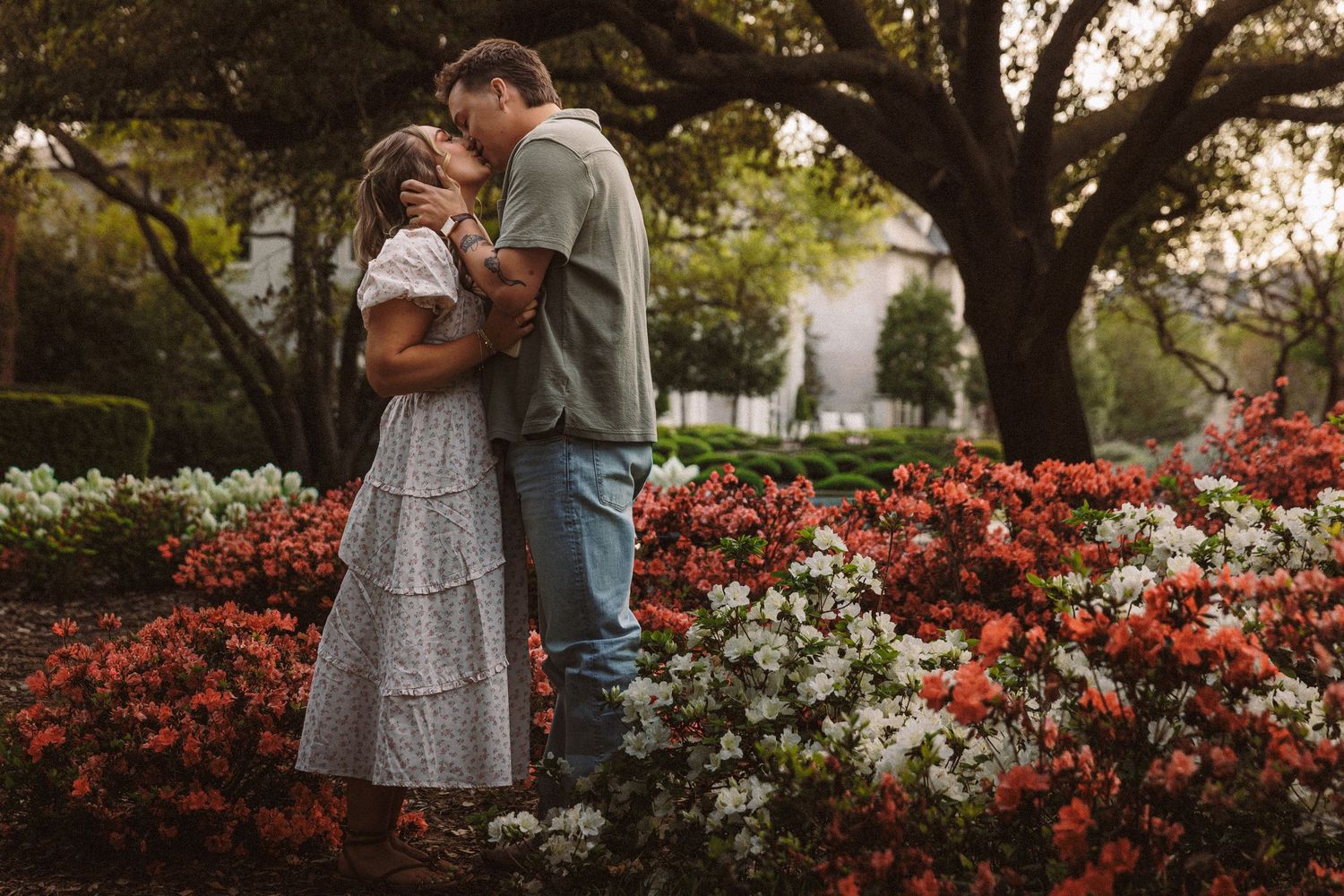 Couple kissing in a garden surrounded by vibrant red and white azalea flowers beneath lush green trees.