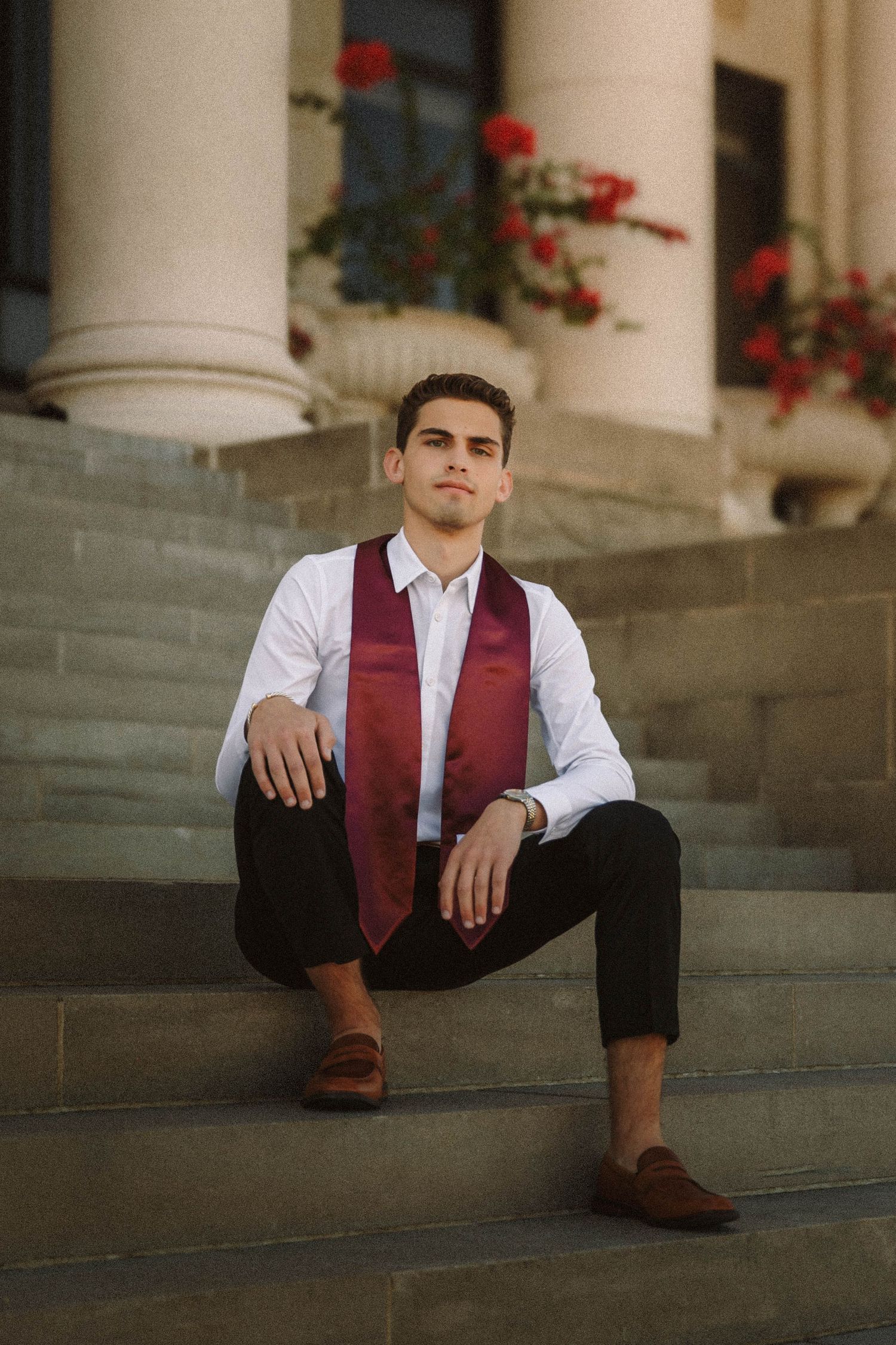 A person in formal attire sits on stone steps with white columns and red flowers in the background.