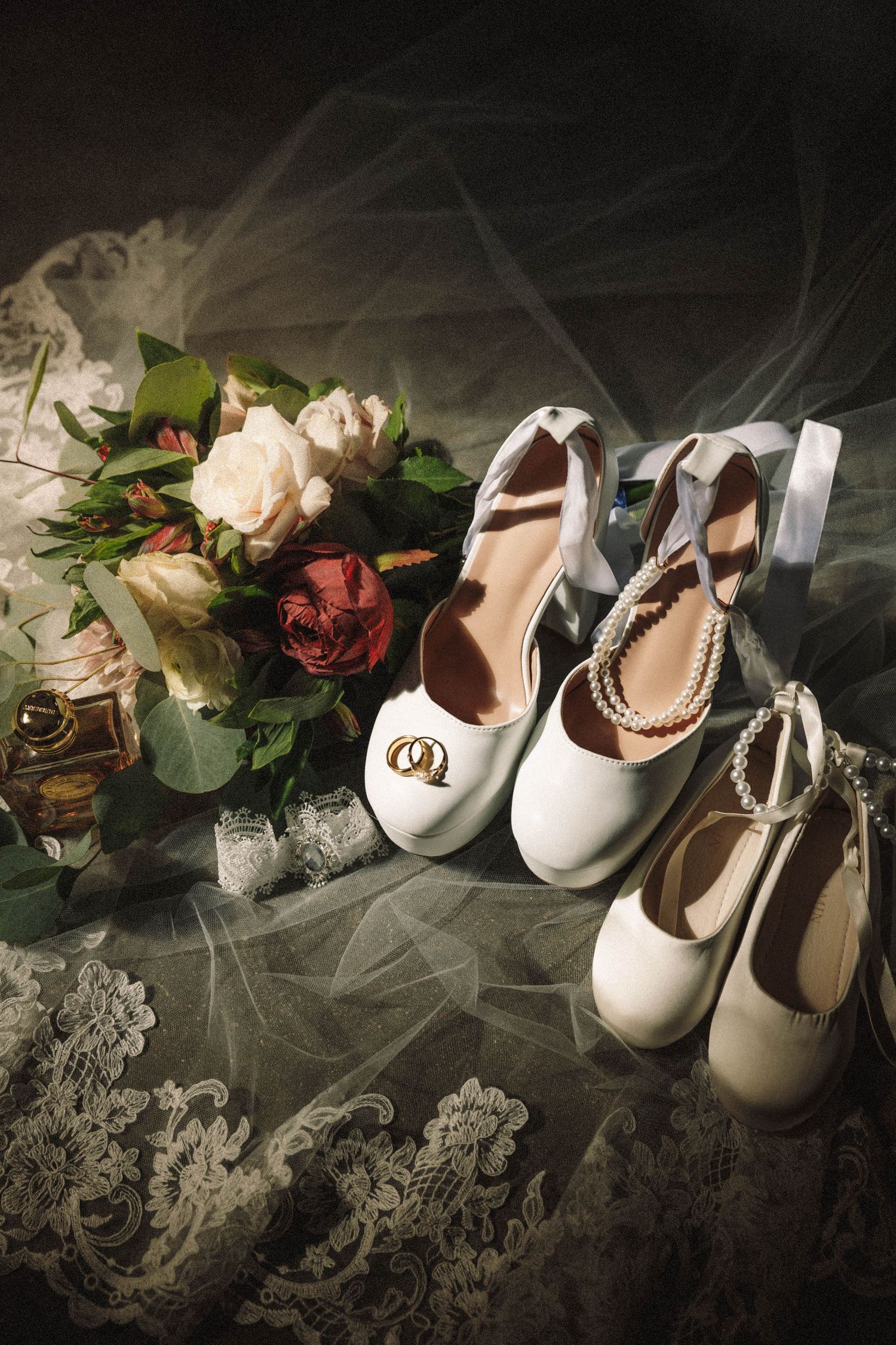 White baby shoes with pearls and lace arranged on dark background with soft lighting and floral details.