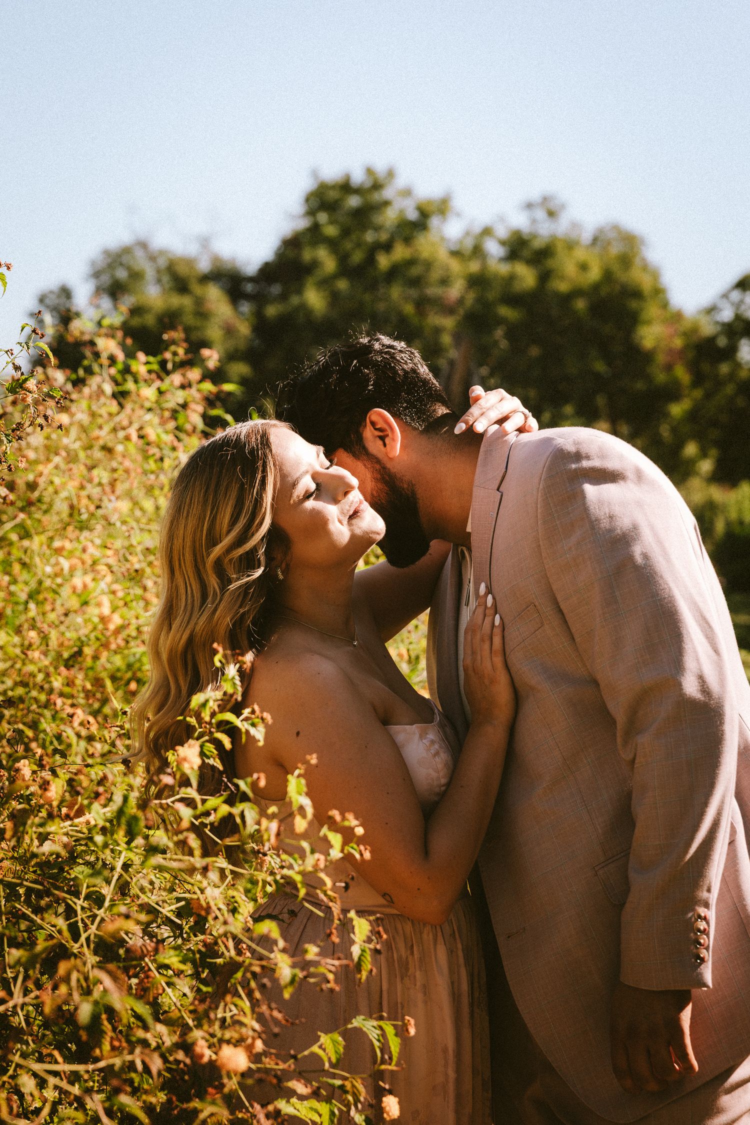 A tender moment between a couple in soft beige attire against a sunlit natural backdrop.