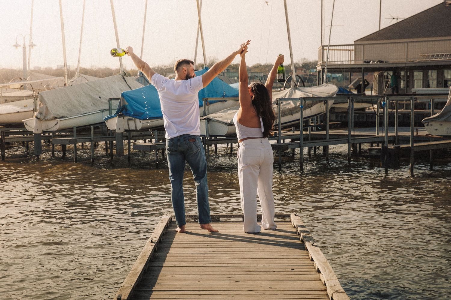 Two people walk hand in hand along a wooden dock by the water.