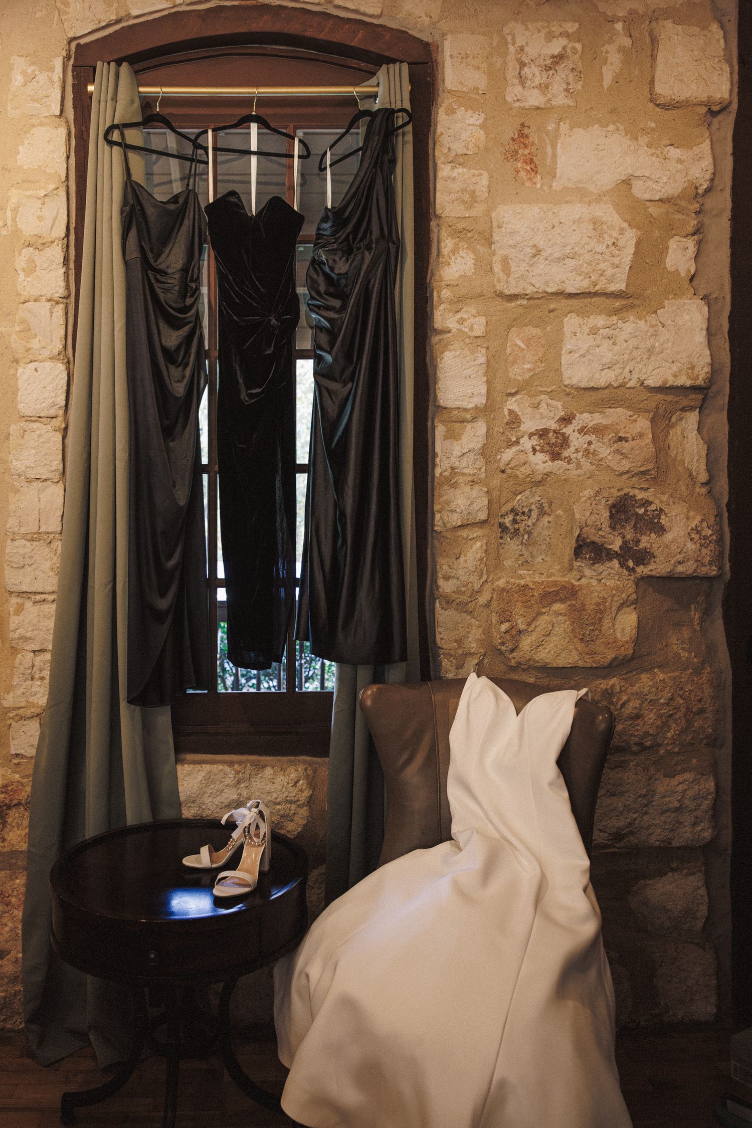 Wedding dress and dark bridesmaid gowns hanging near stone wall window with bridal heels on table.