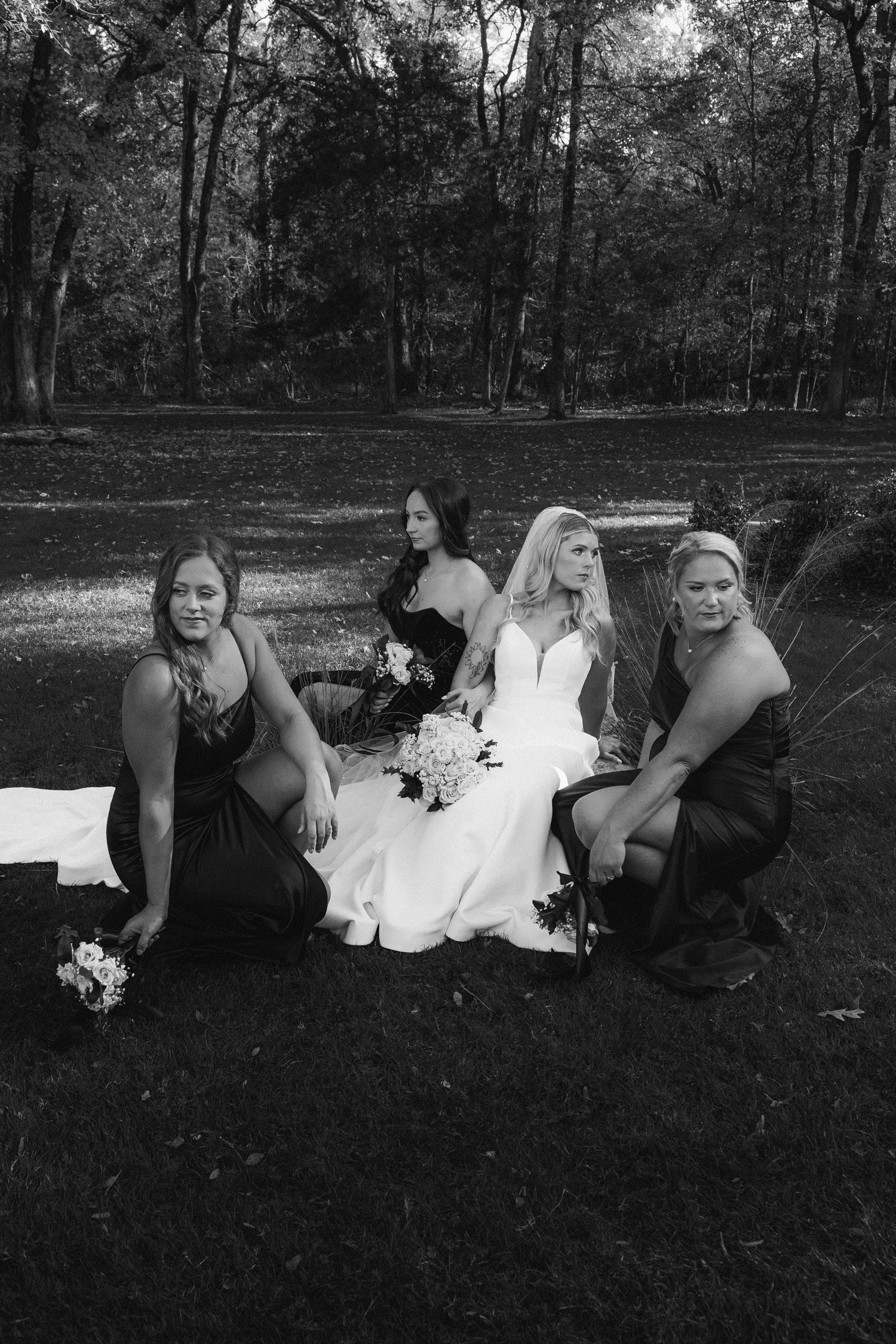 Black and white photo of bride in white gown seated outdoors surrounded by three bridesmaids in dark dresses.