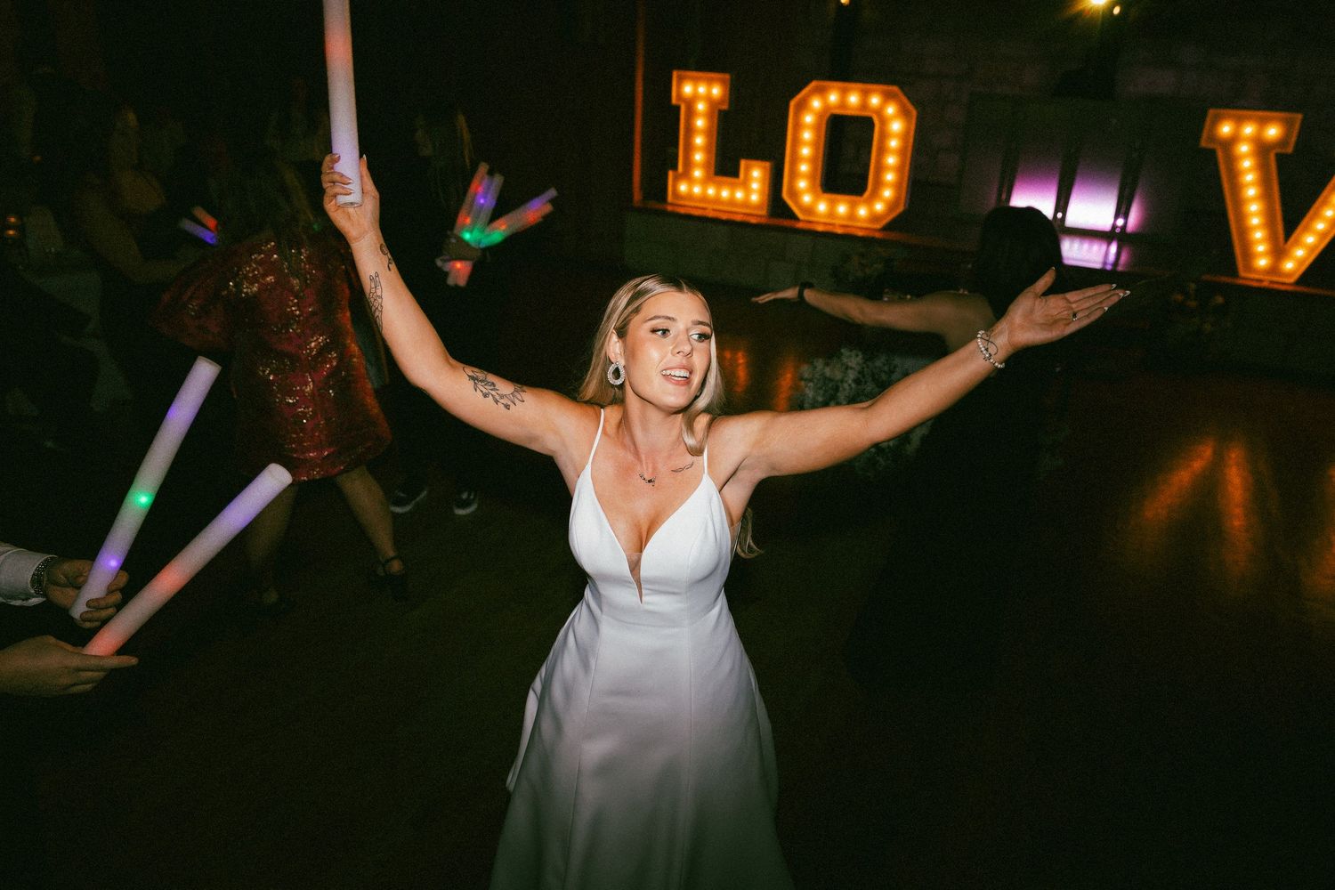 Joyful bride dancing with arms raised near illuminated LOVE marquee letters at wedding reception.