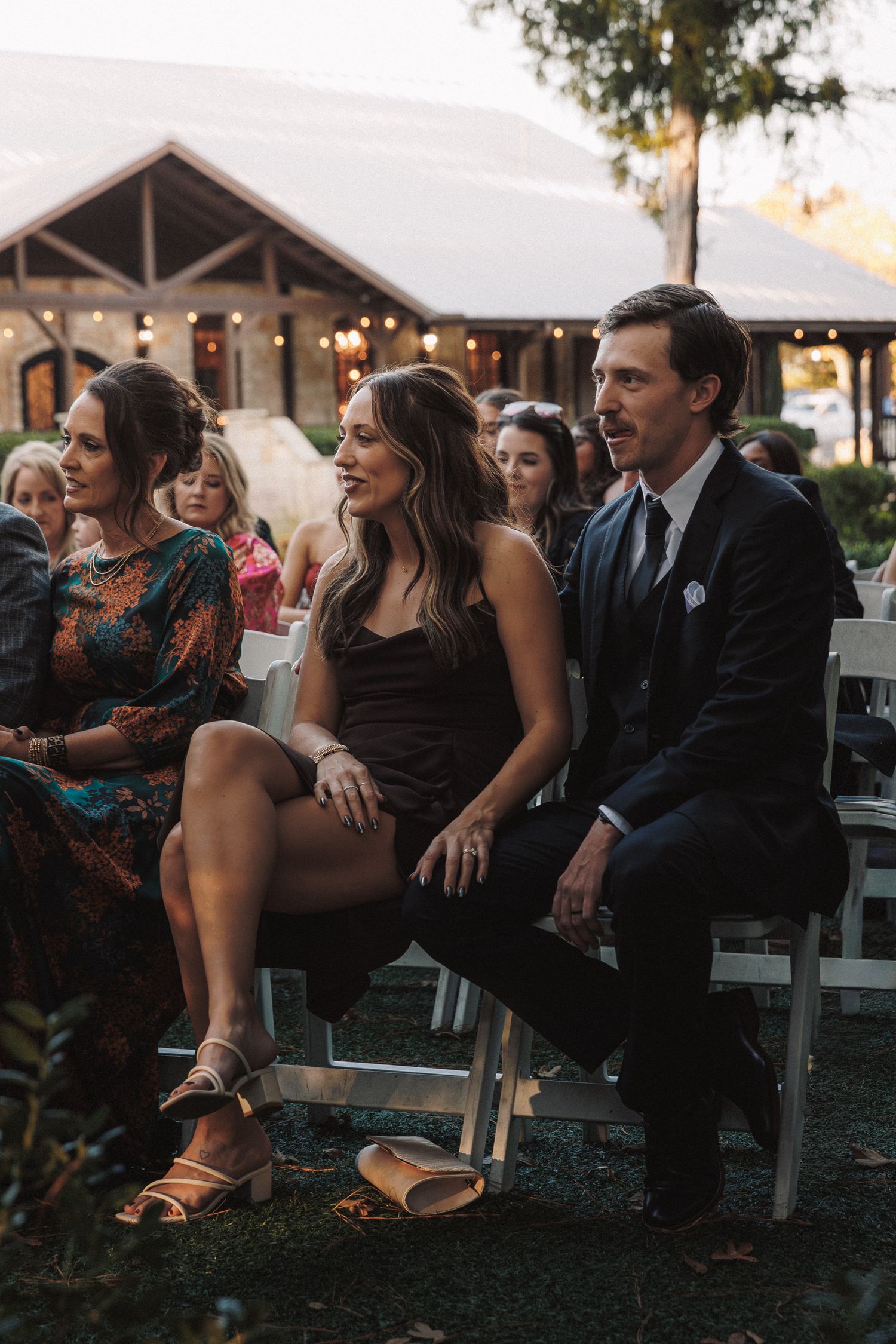 Couple in formal attire seated at outdoor wedding reception, man in navy suit and woman in black dress smiling.