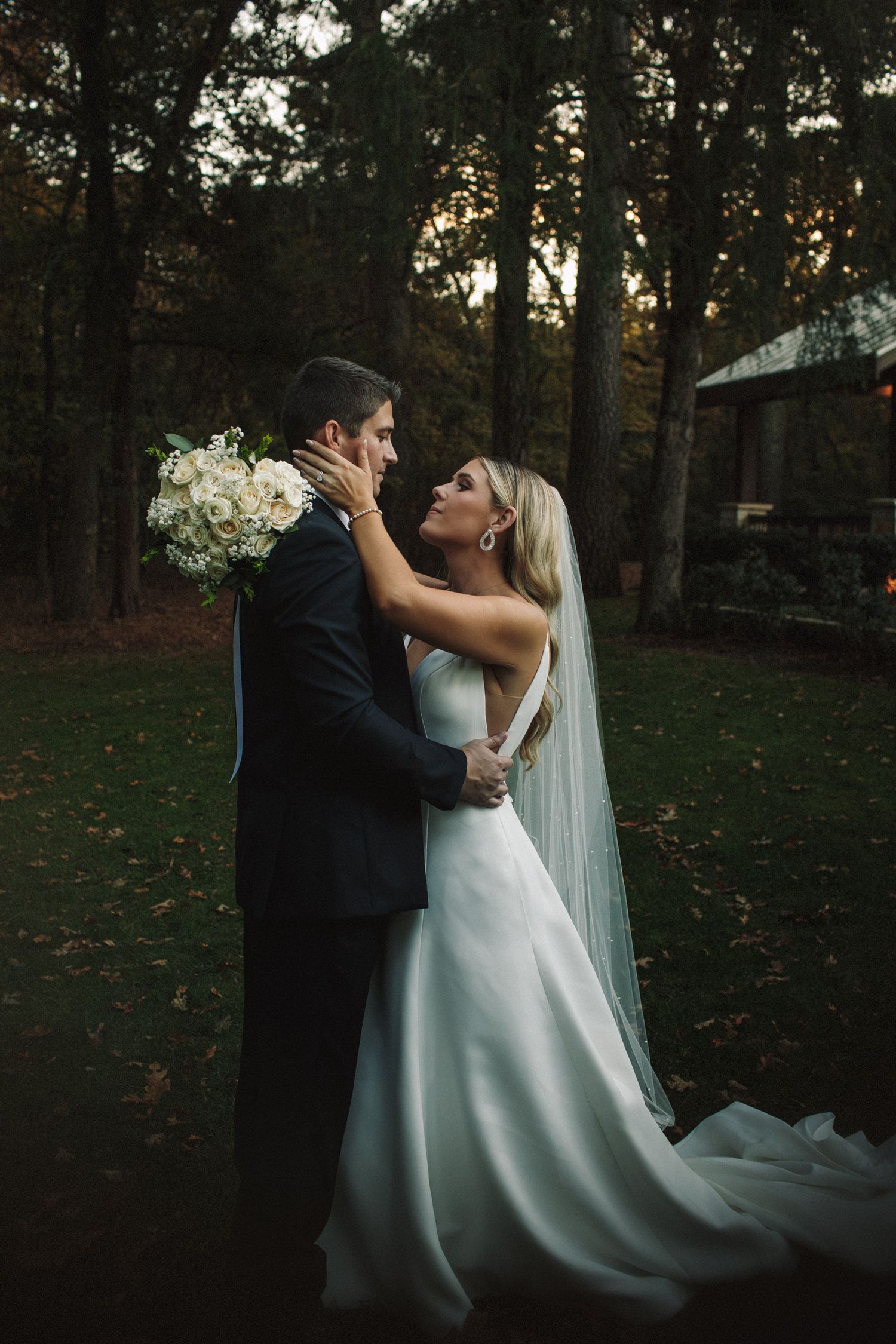 Bride and groom share a romantic embrace at dusk surrounded by tall trees.