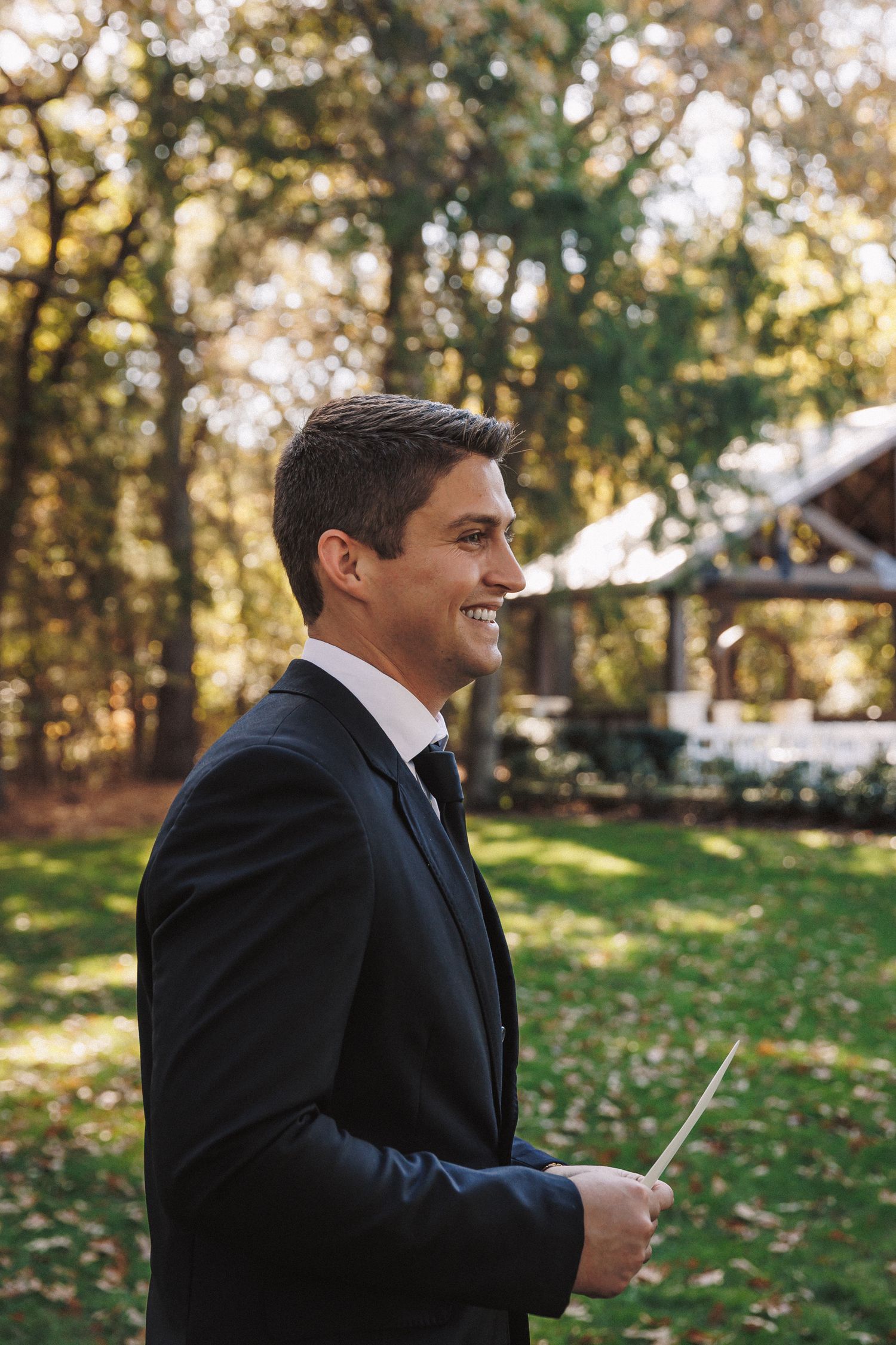 Smiling groom in black tuxedo holding card outdoors with autumn trees and wedding pavilion behind him.