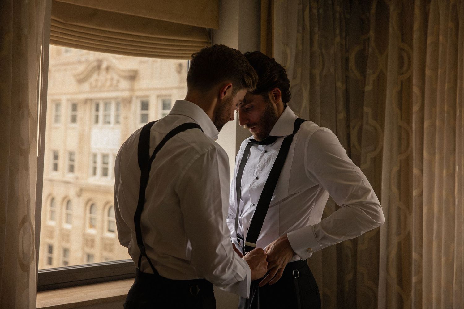Two grooms in white shirts and suspenders sharing an intimate moment by a hotel window.
