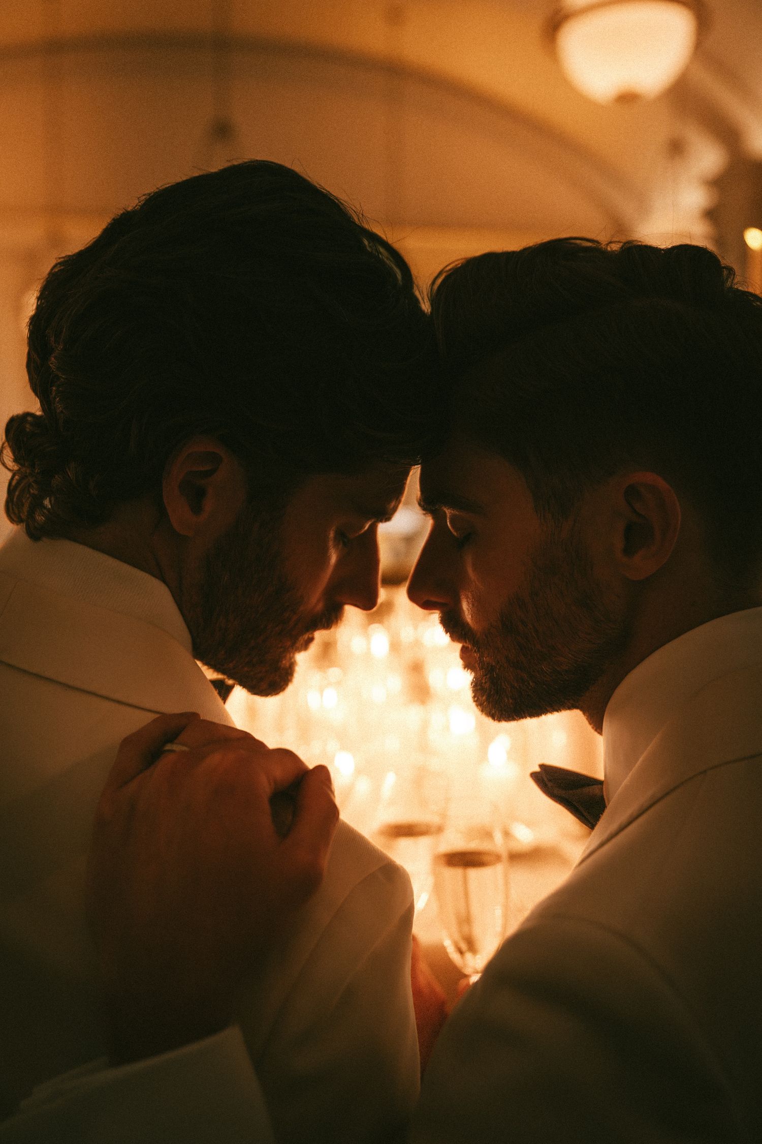 Two grooms in white suits share an intimate forehead touch by candlelight at their wedding reception.
