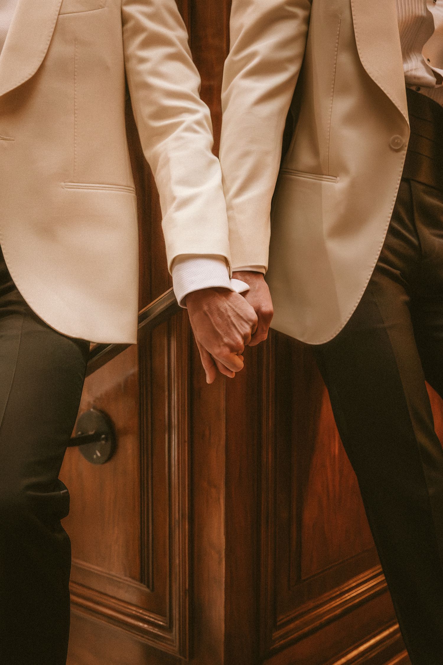 Close-up of two grooms holding hands in matching cream tuxedo jackets at wedding.