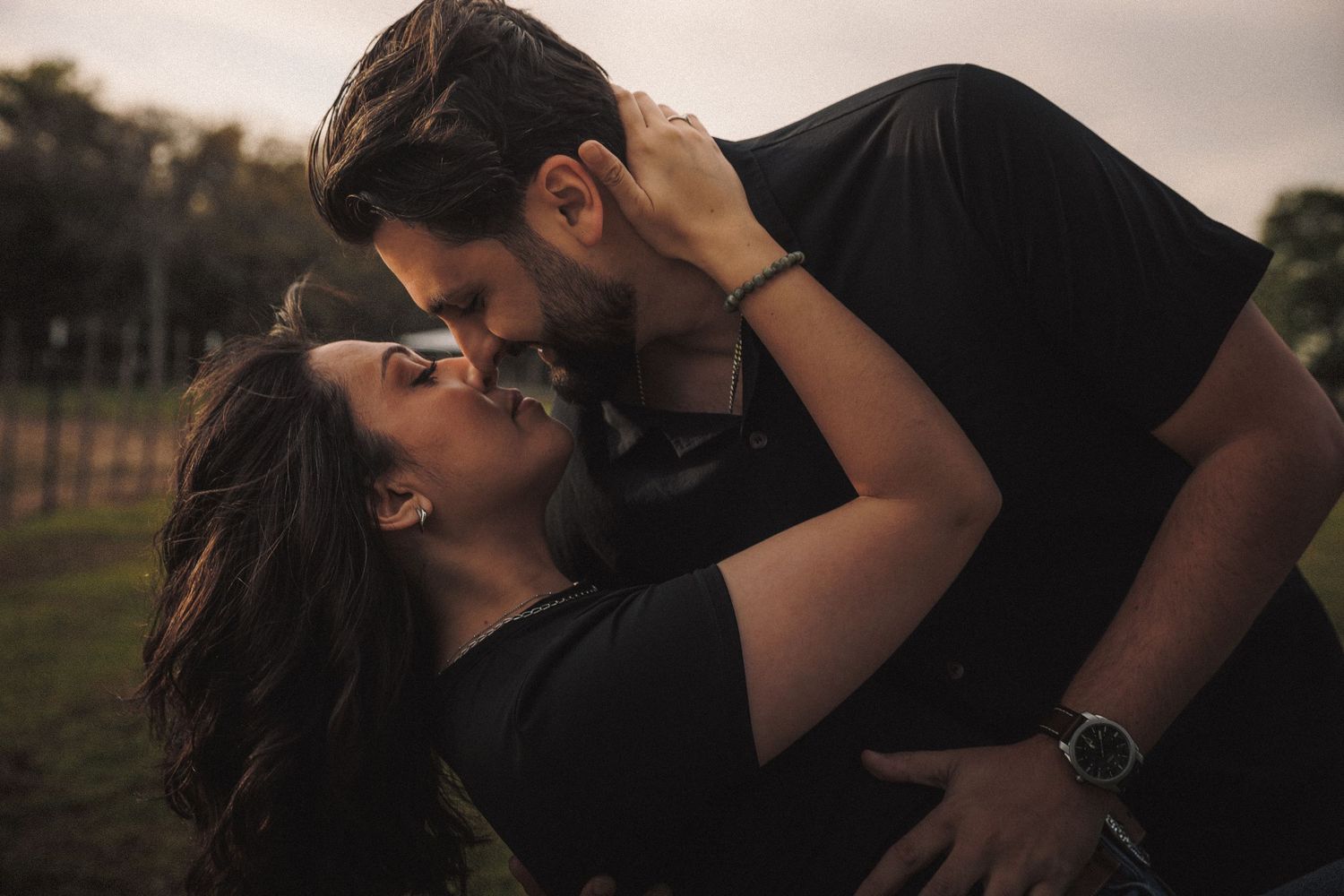 Romantic couple in black outfits sharing a passionate kiss outdoors at dusk.