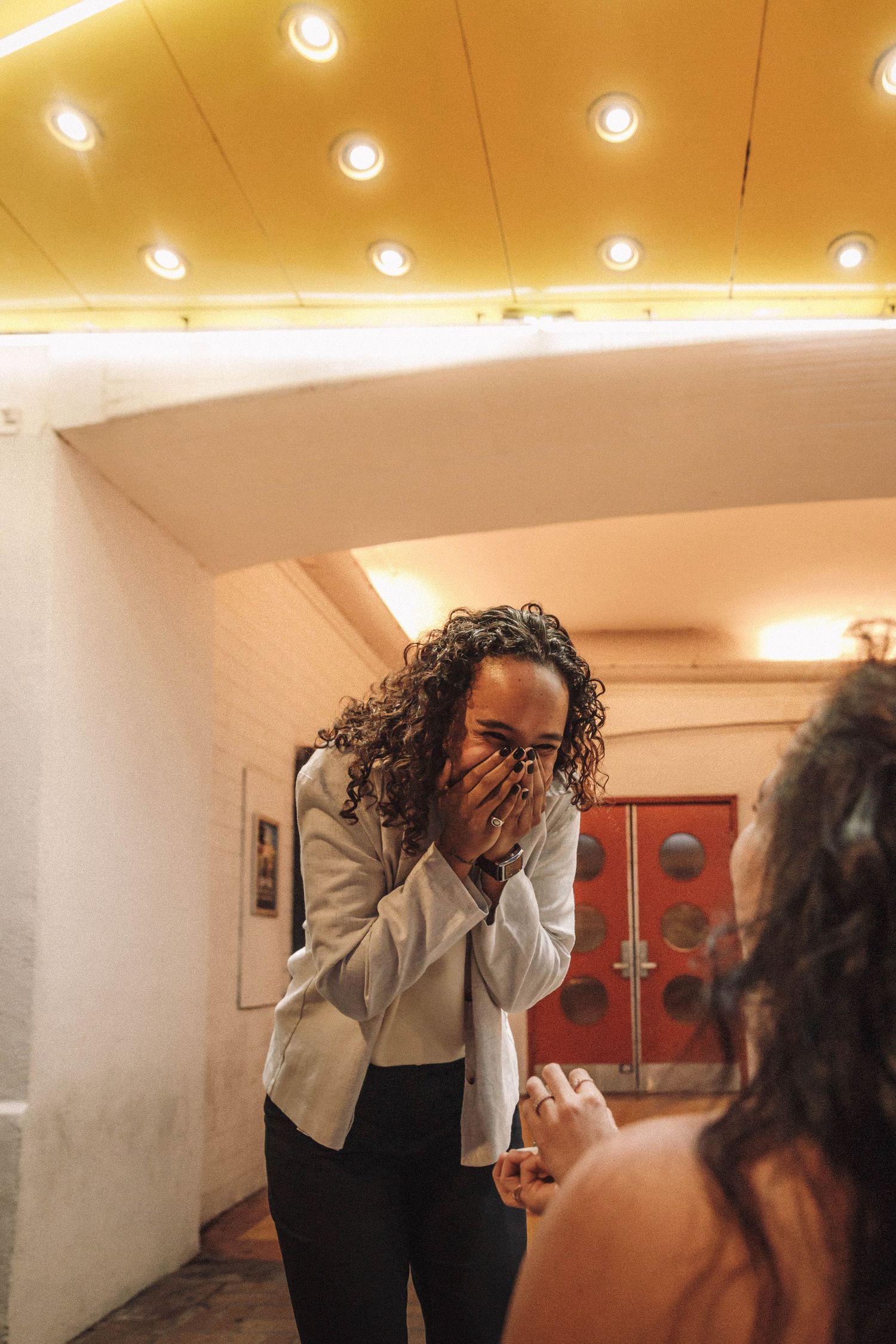 Woman covers mouth in emotional surprise during a marriage proposal inside a theater.
