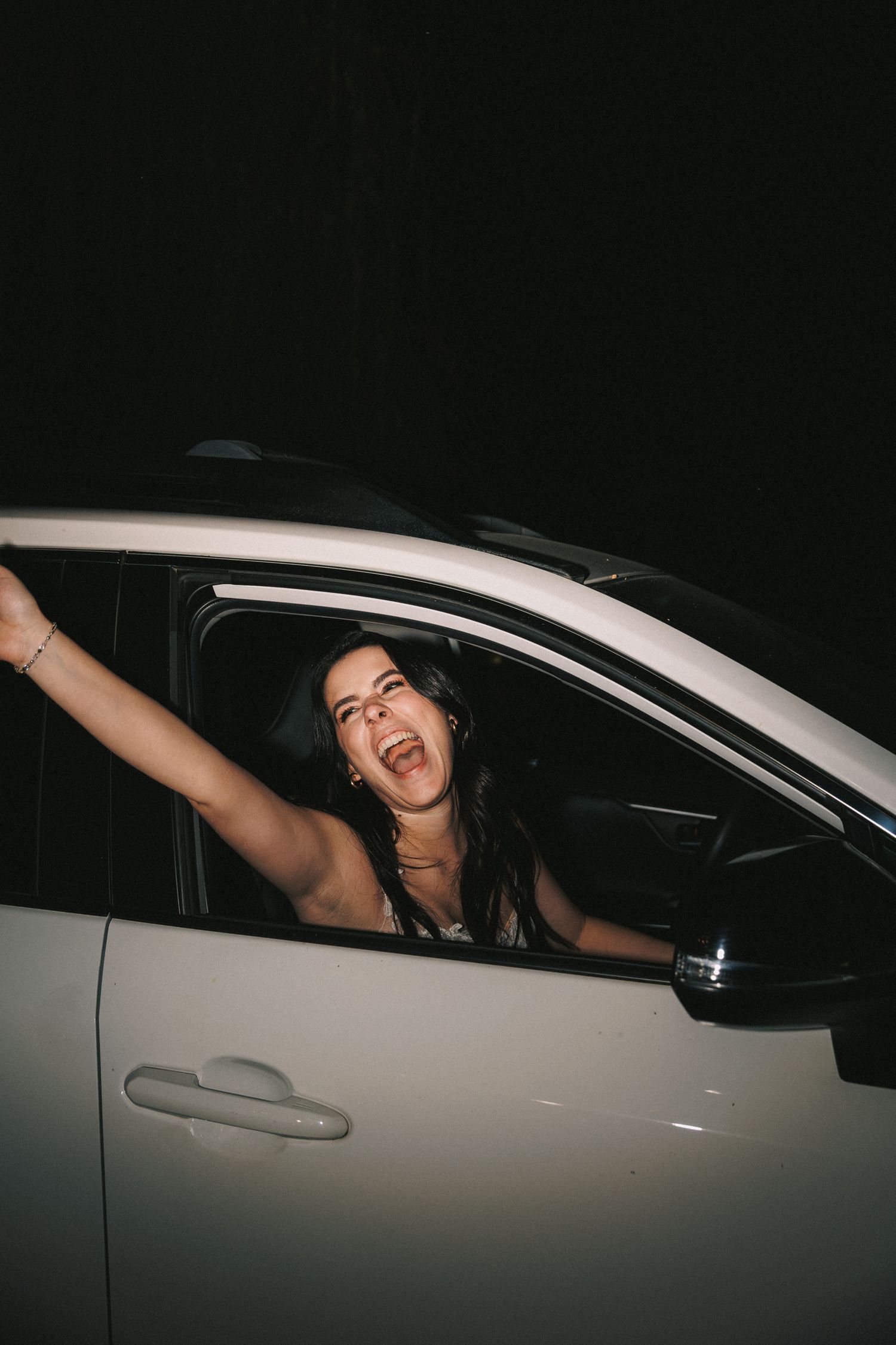 Joyful bride leans out of white car window celebrating after her wedding reception at night.