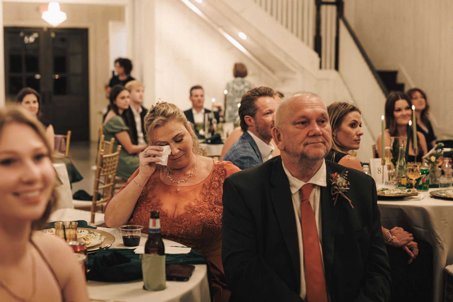 Wedding guests seated at reception tables, with emotional woman dabbing tears during speeches.