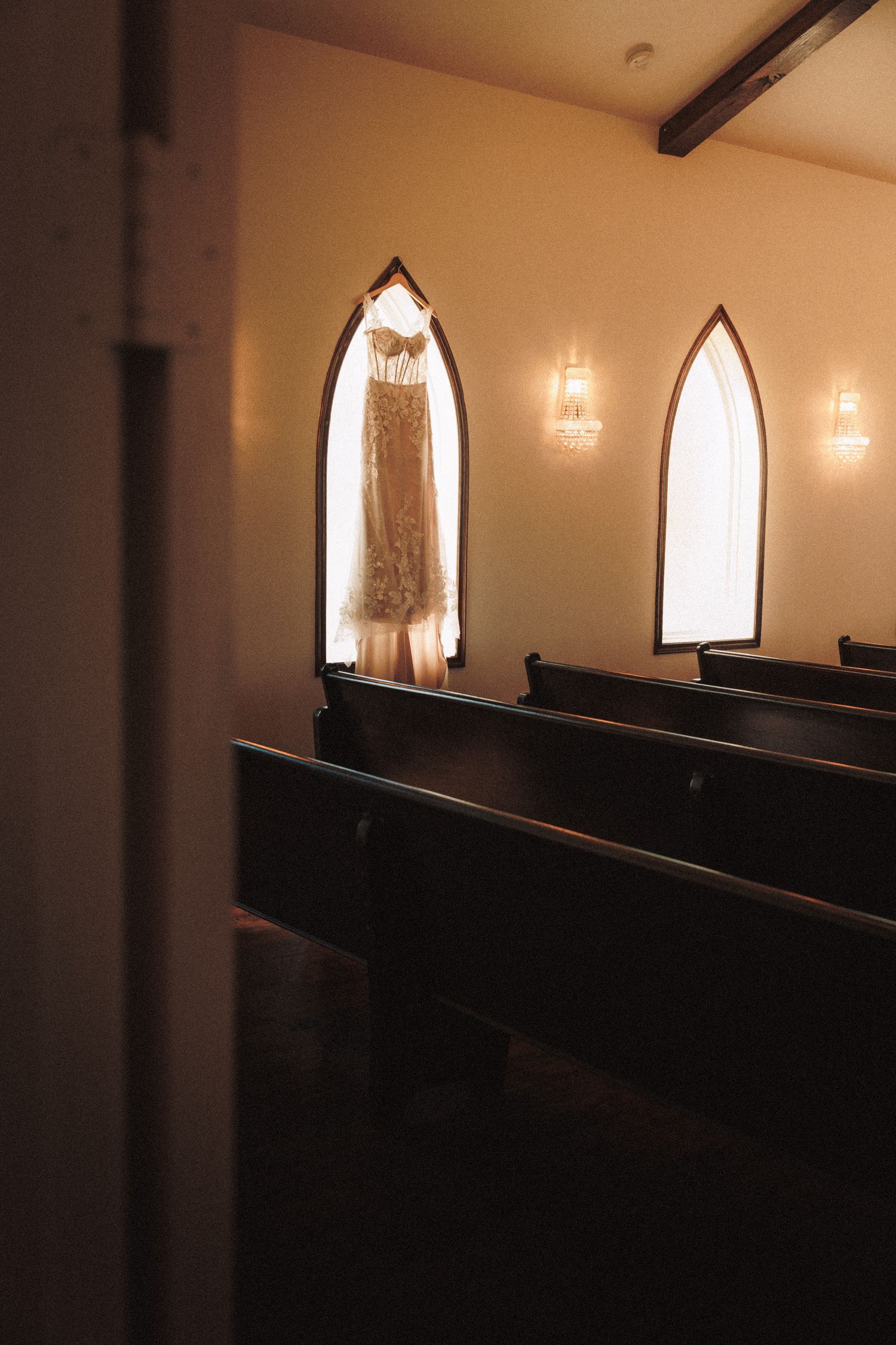 Lace wedding dress hanging in arched chapel window with wooden pews in foreground.