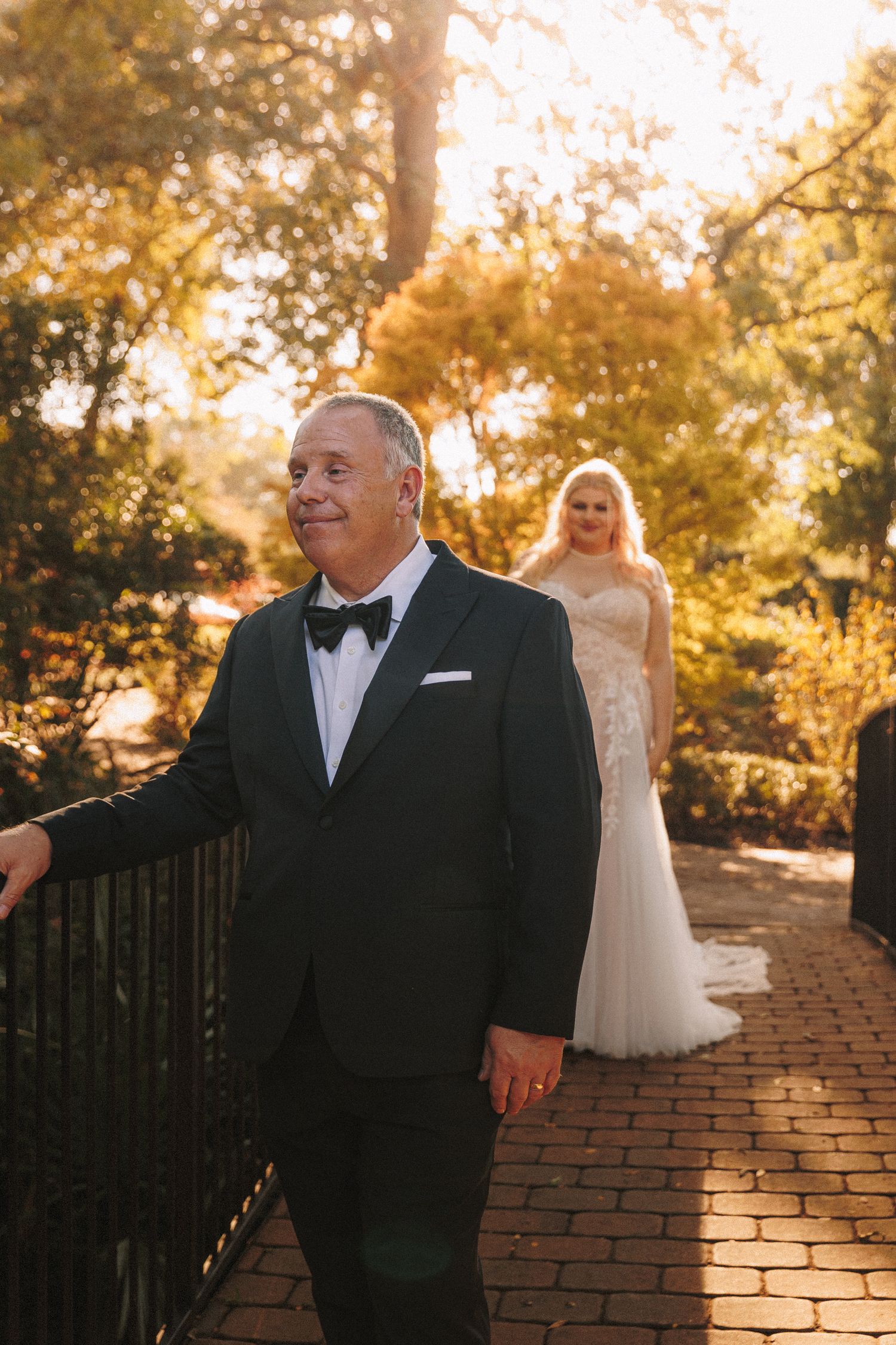 Bride in lace wedding gown approaches man in black tuxedo on a brick path in a sunlit garden.