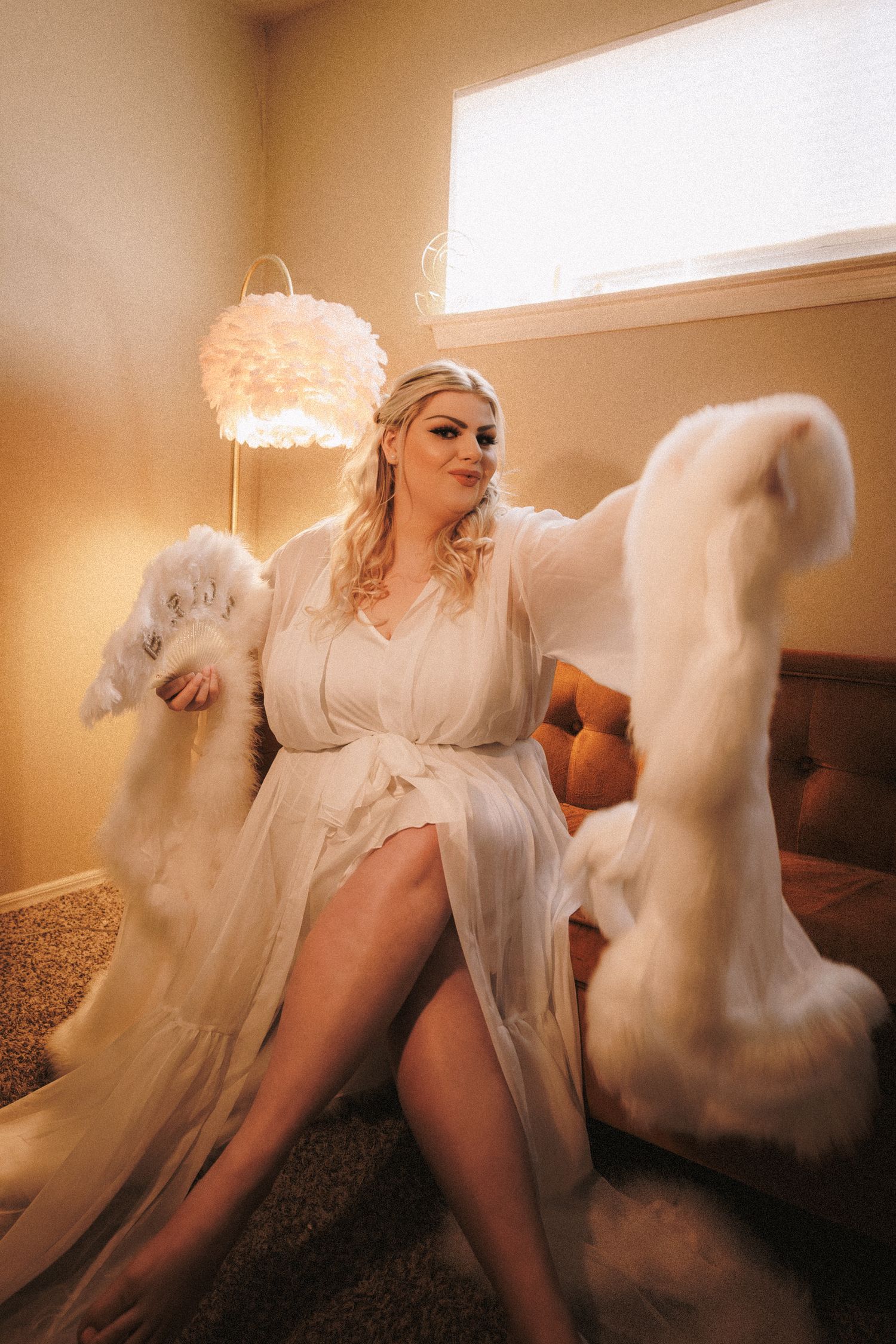 Bride in white sheer feather-trimmed robe sitting elegantly on a couch in a warmly lit room.
