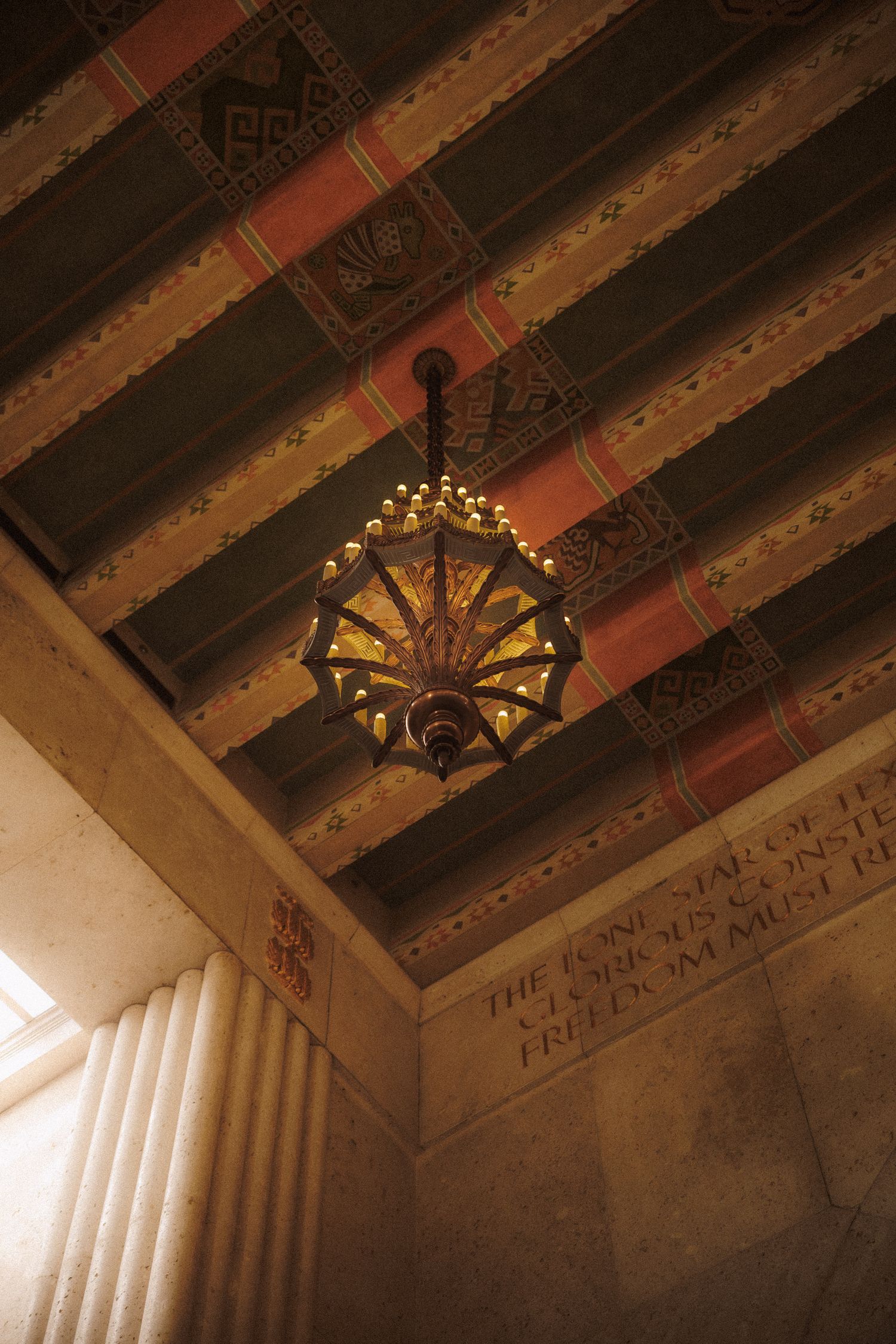 Ornate vintage chandelier hanging from decorative painted ceiling with engraved stone columns.