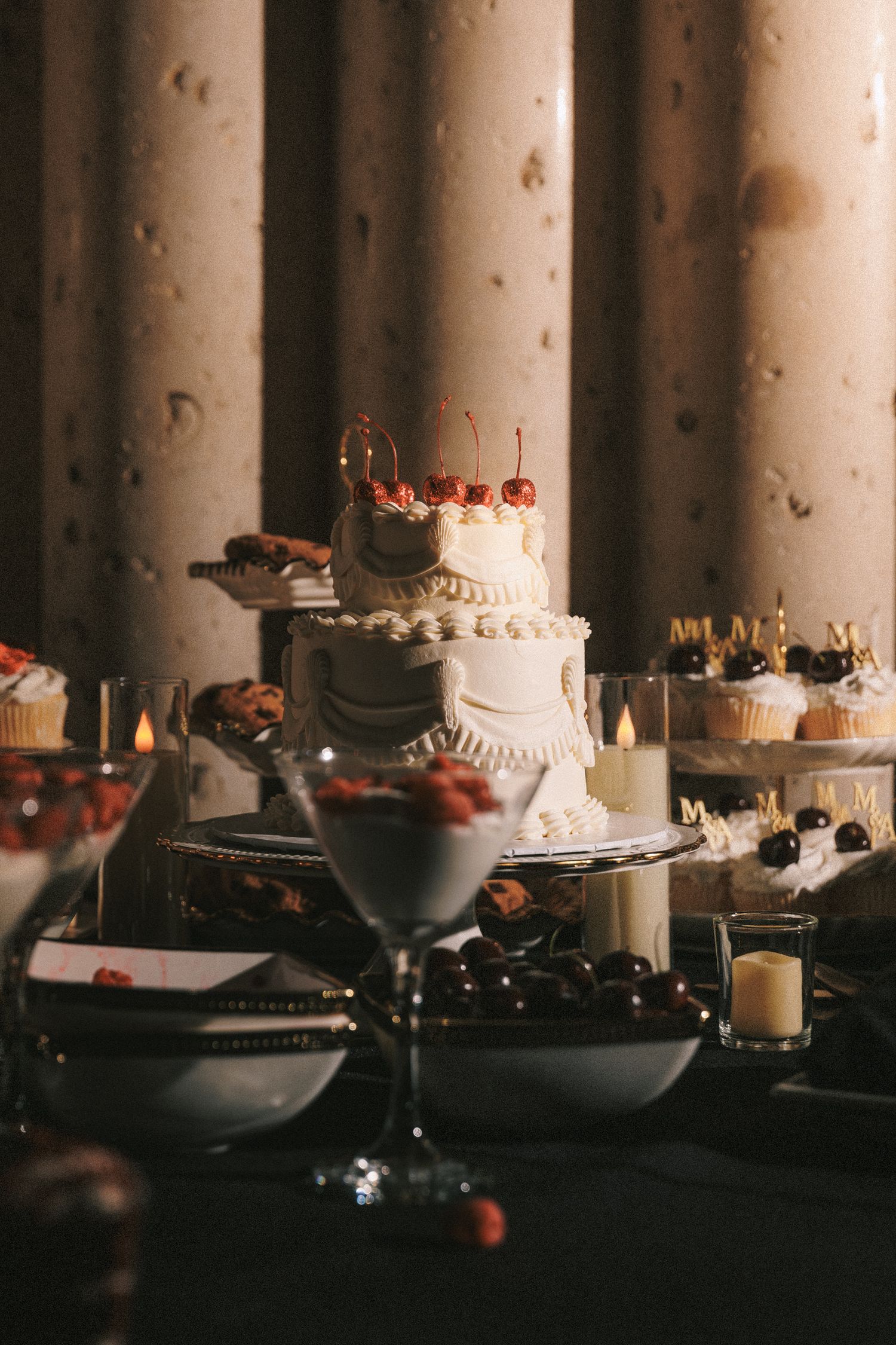 Elegant white two-tier wedding cake topped with cherries surrounded by desserts and candles.