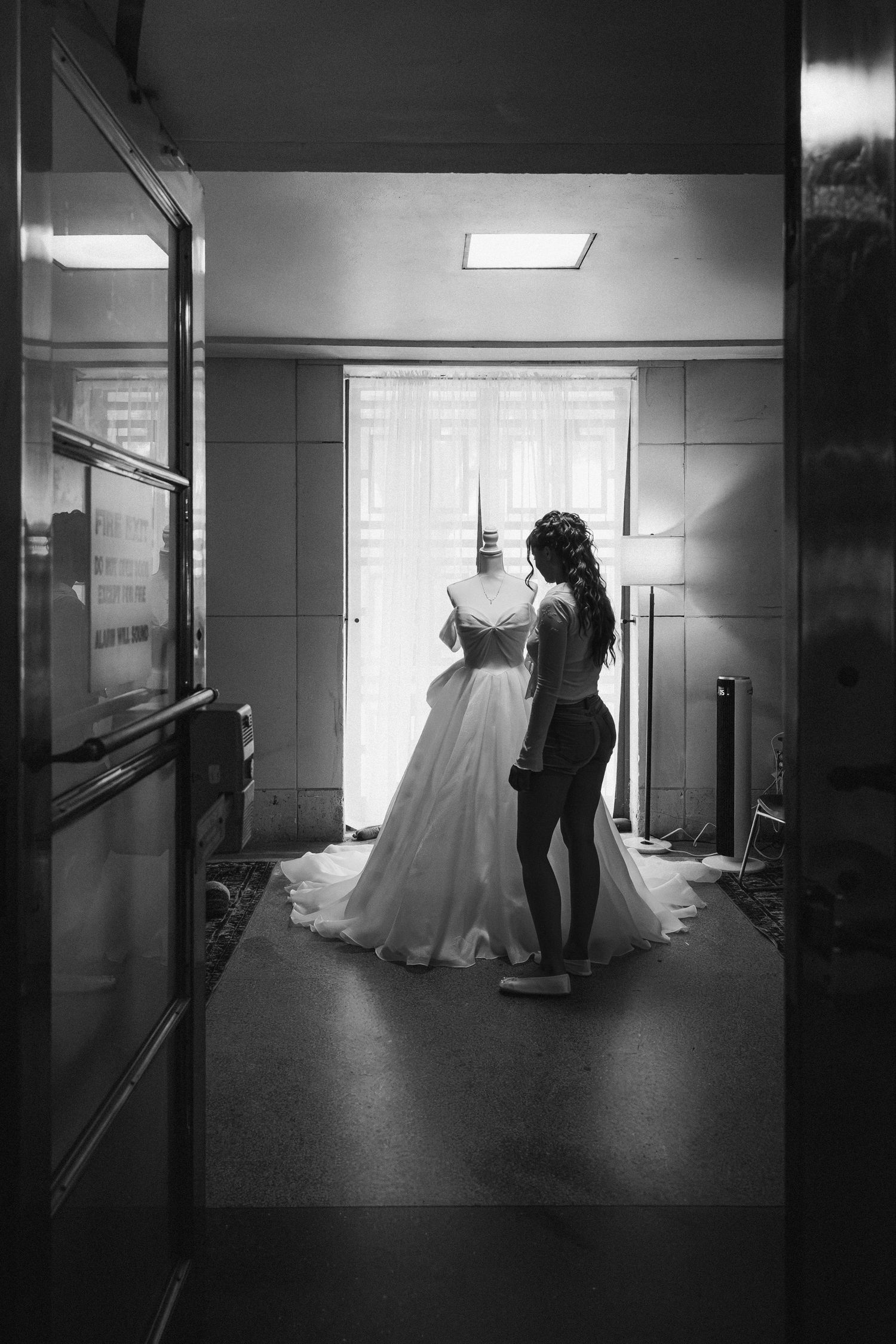 Black and white photo of bride adjusting wedding gown on mannequin in elegant hallway.
