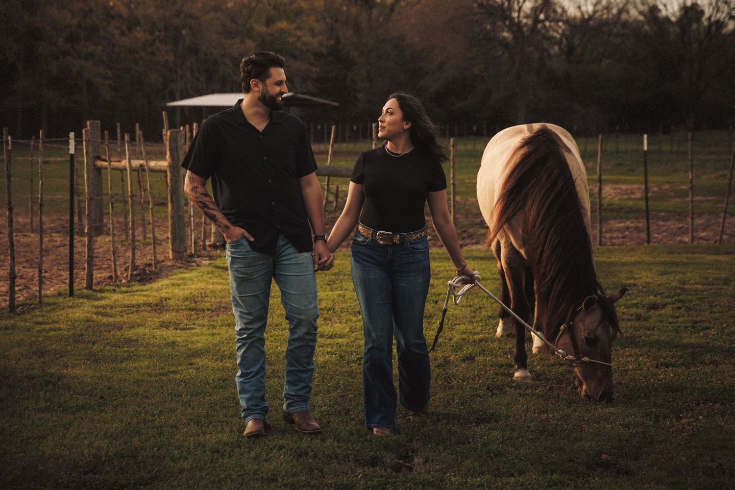 Couple holding hands and walking near a grazing horse on a farm at golden hour.