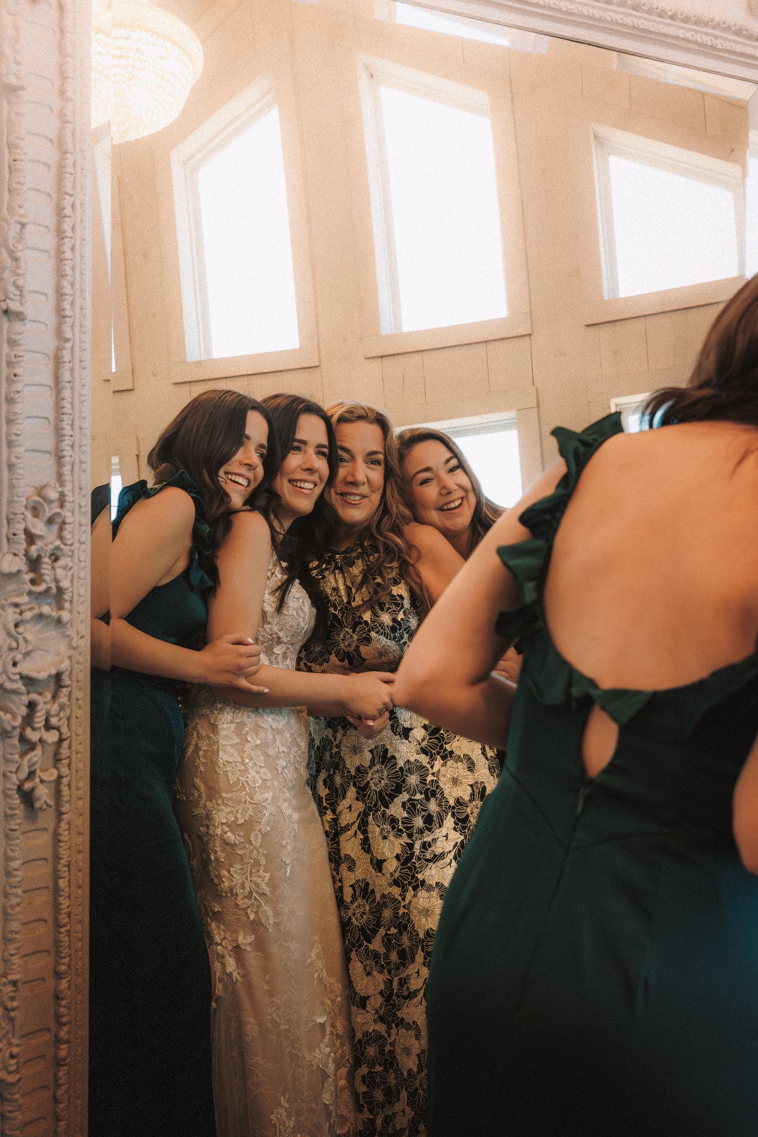 Bride in lace gown hugging bridesmaids in emerald green dresses reflected in ornate mirror.