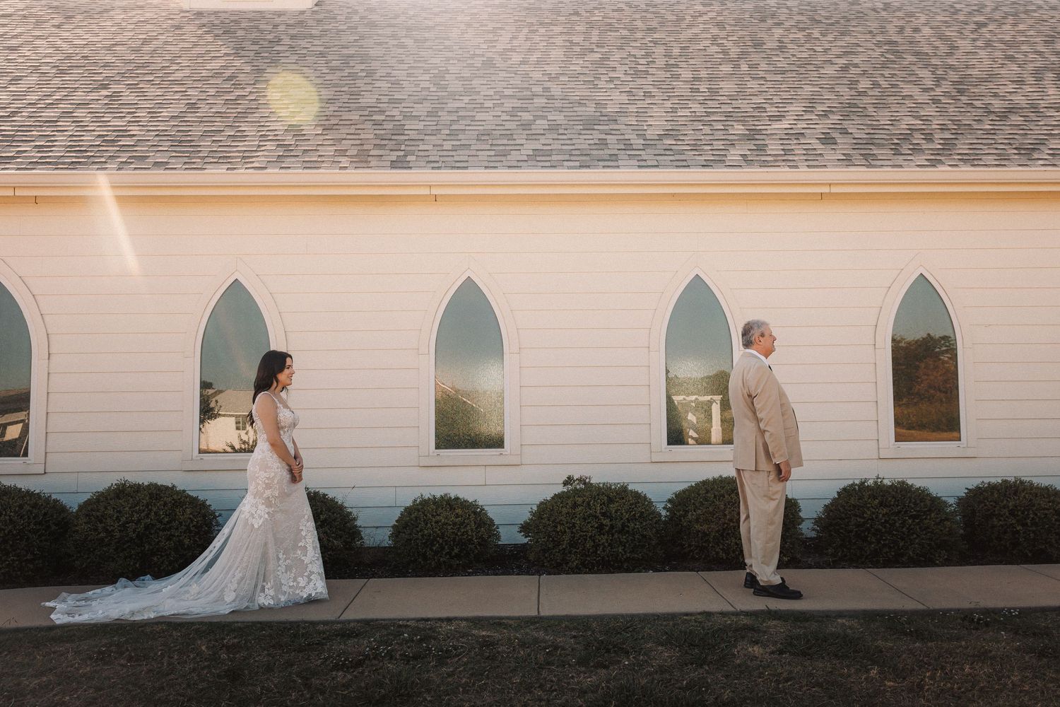 Bride and groom standing apart for a first look outside a white chapel with arched windows.